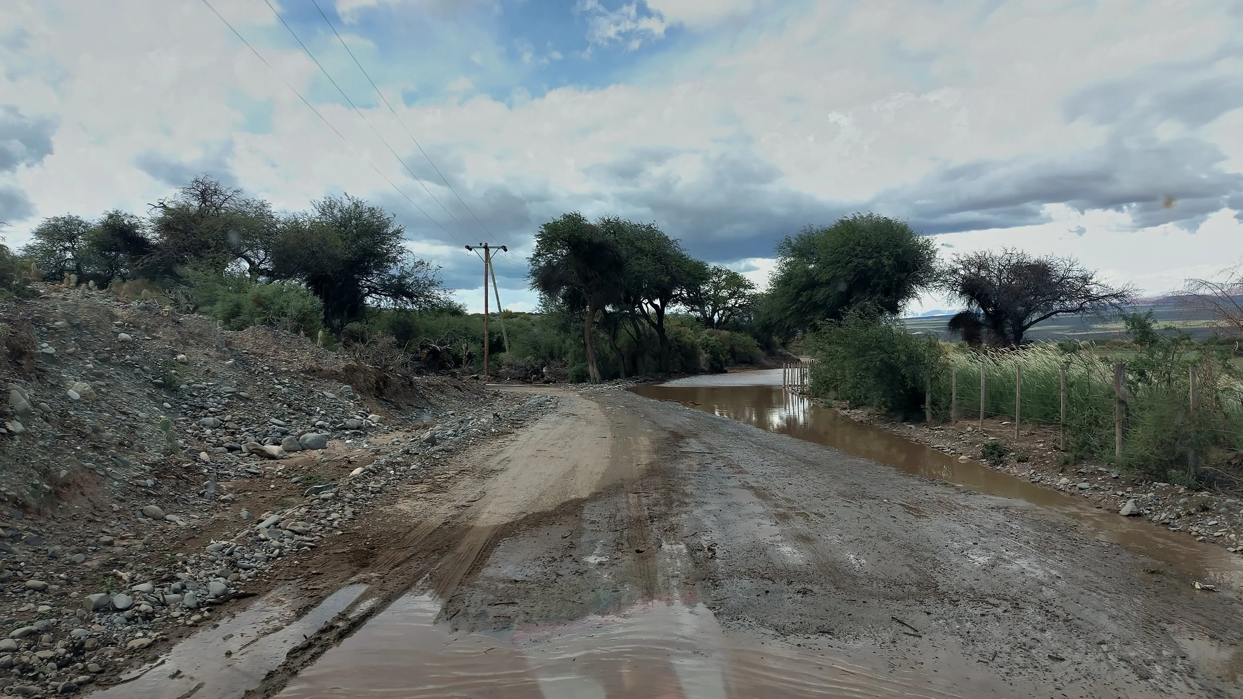 Eine unbefestigte, schlammige Straße verläuft durch eine ländliche Gegend, flankiert von Bäumen und einem Zaun auf der rechten Seite, mit einem kleinen Wasserlauf auf der Straße, helle Wolken am Himmel.
