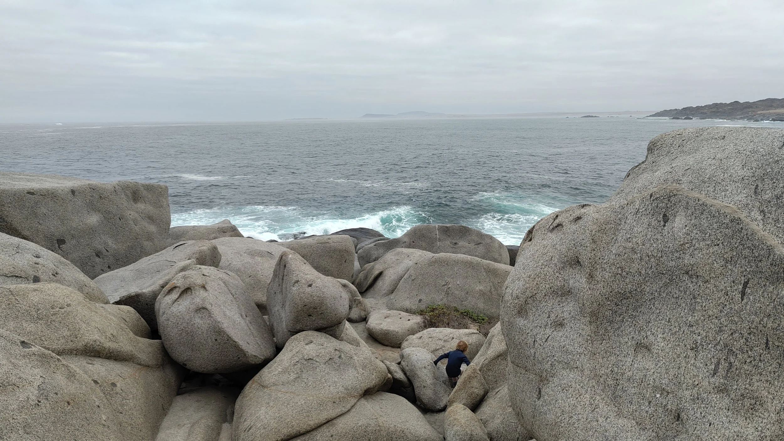Felsen am Meer mit einem Kind, das zwischen den Steinen spielt, unter bewölktem Himmel.