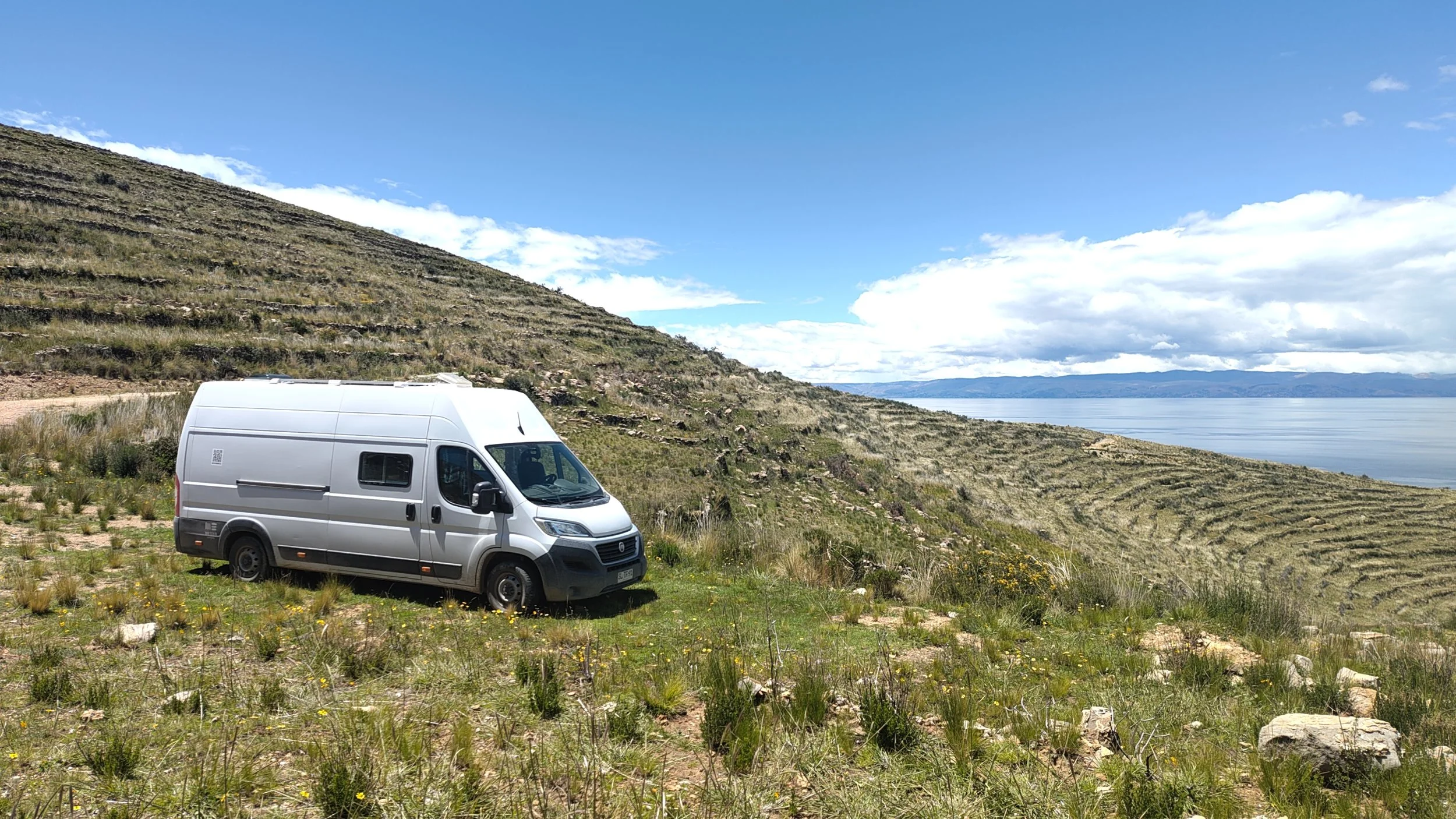 Ein weißer Lieferwagen steht auf einer Wiese vor einer hügeligen Landschaft mit Gräsern und kleinen Blumen, im Hintergrund ein See und bewölkter Himmel.