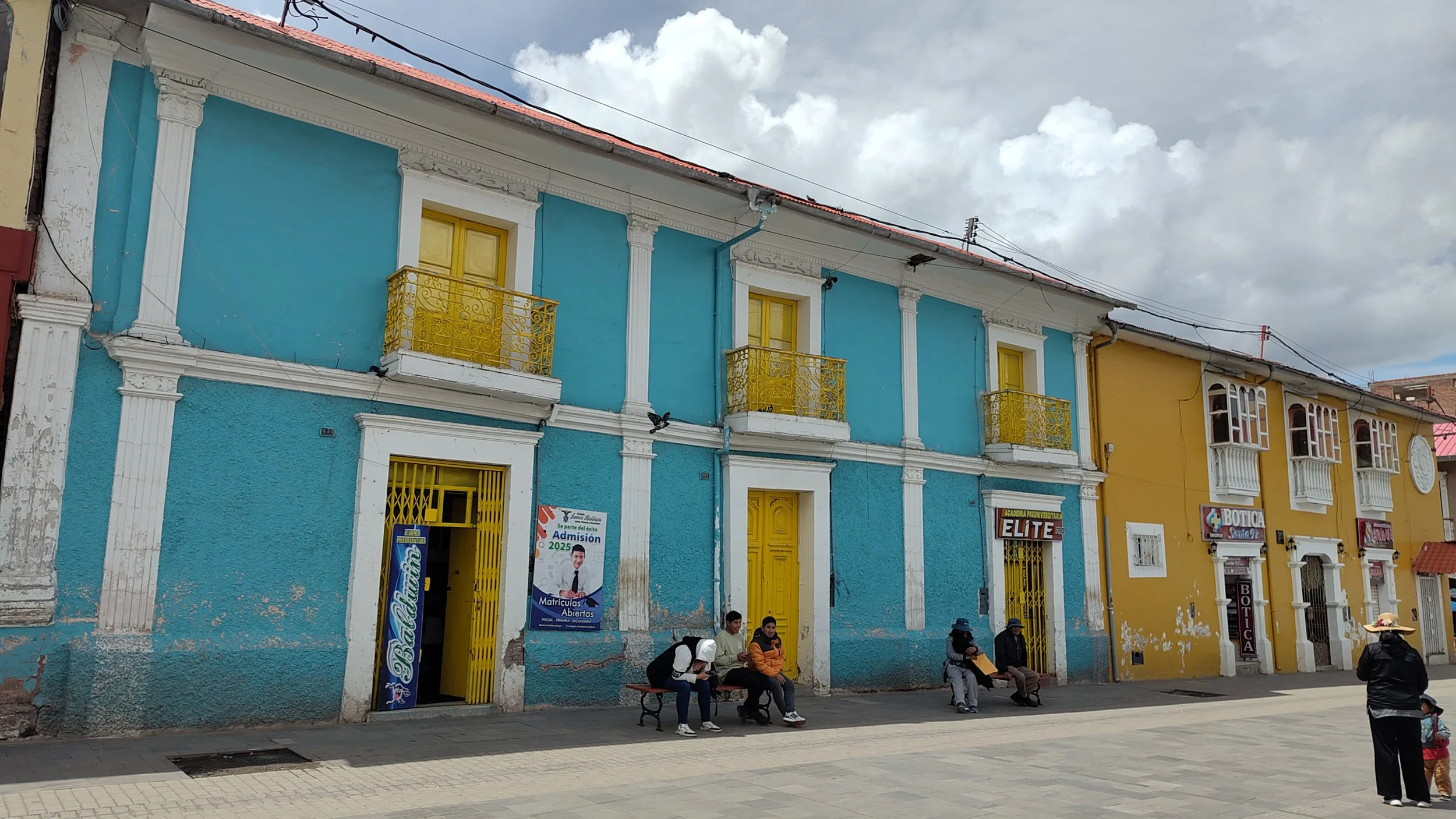Bunte zweistöckige Gebäude in Blau und Gelb, mit Balkonen und Fenster, Menschen sitzen und stehen vor dem Gebäude, über dem Gebäude sind Wolken am Himmel.