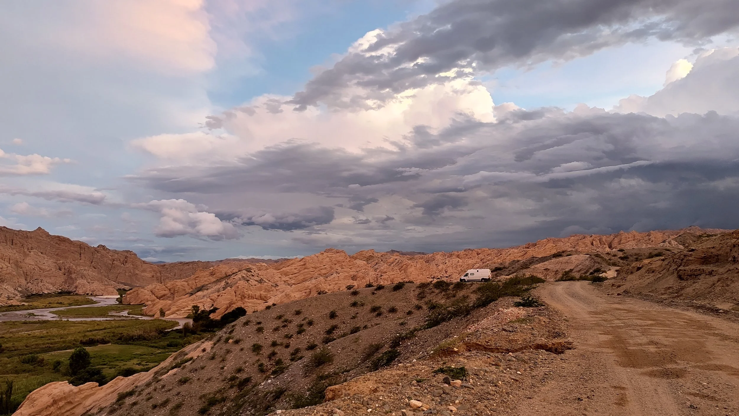 Ein weißer Camper steht auf einer staubigen Straße in einer Wüstenlandschaft mit orangefarbenen Felsen und einigen grünen Pflanzen, im Hintergrund sind Hügel und eine bewölkte Himmel zu sehen.