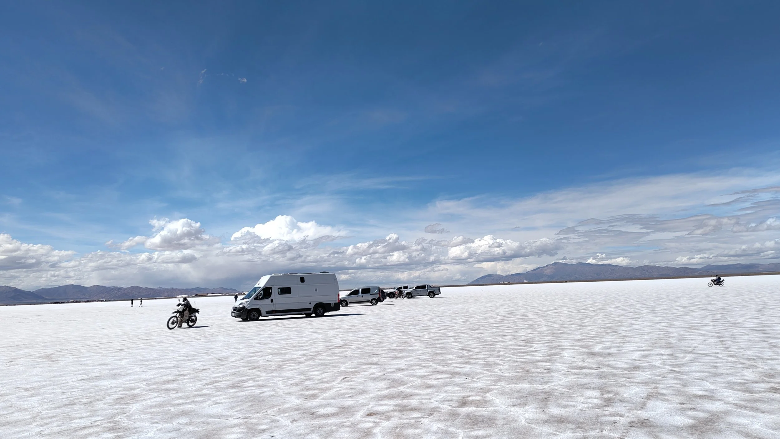Weißer Salzsee mit mehreren Fahrzeugen und Motorrädern, im Hintergrund Berge und ein blauer Himmel mit Wolken.