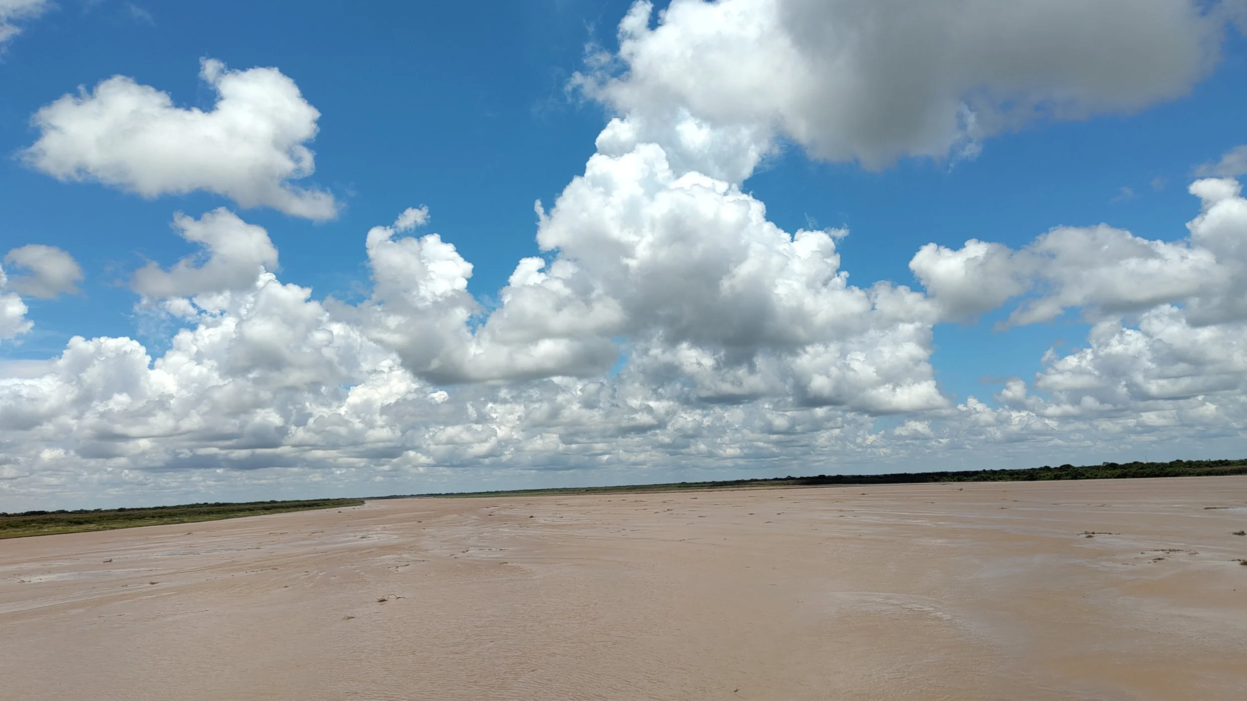 Weite Ebene mit sandigem Boden, grüne Vegetation am Horizont und ein blauer Himmel mit weißen Wolken.
