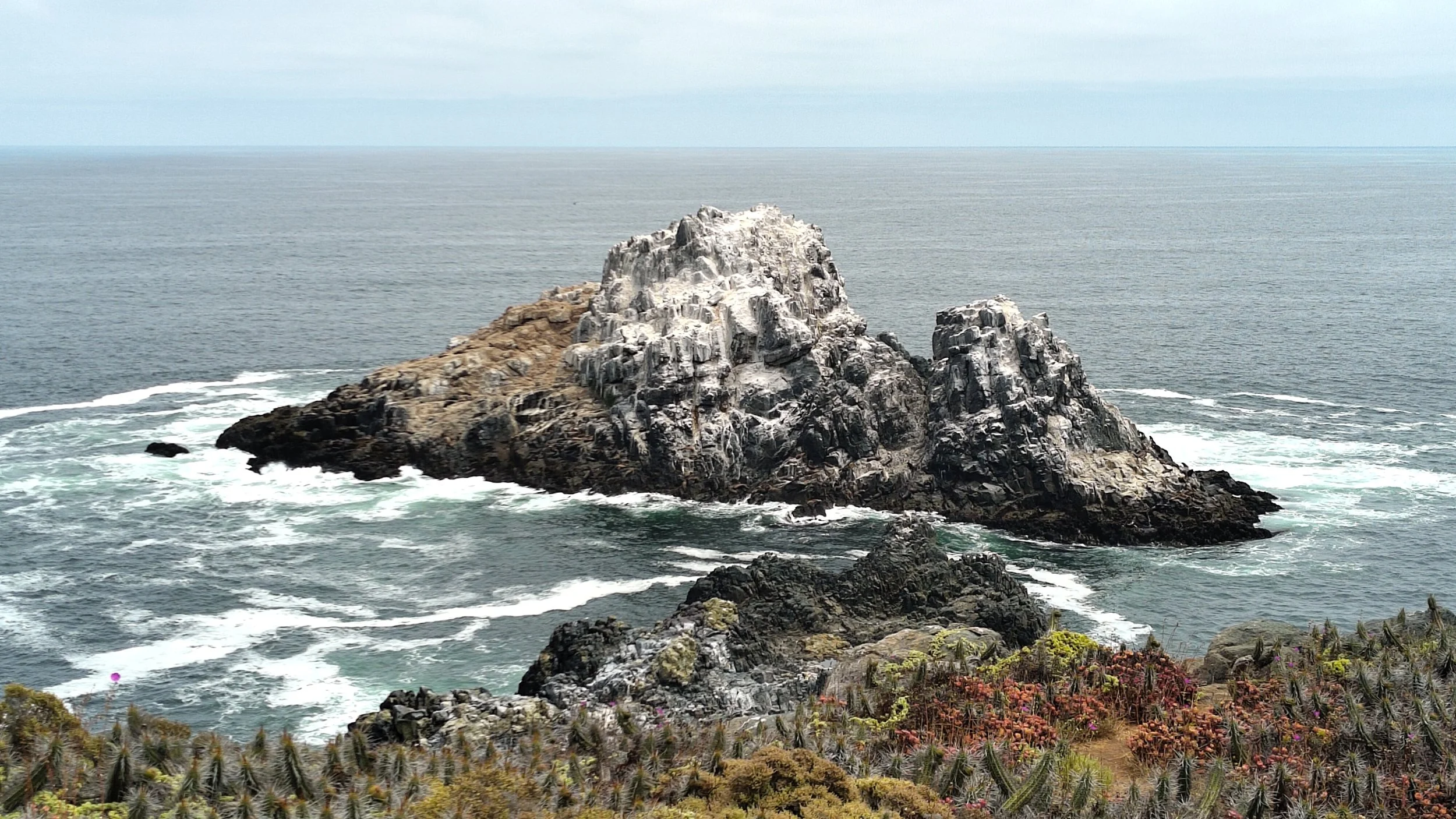 Eine felsige Insel im Meer, umgeben von Wasser mit Wellen, im Hintergrund ein Himmel mit Wolken.