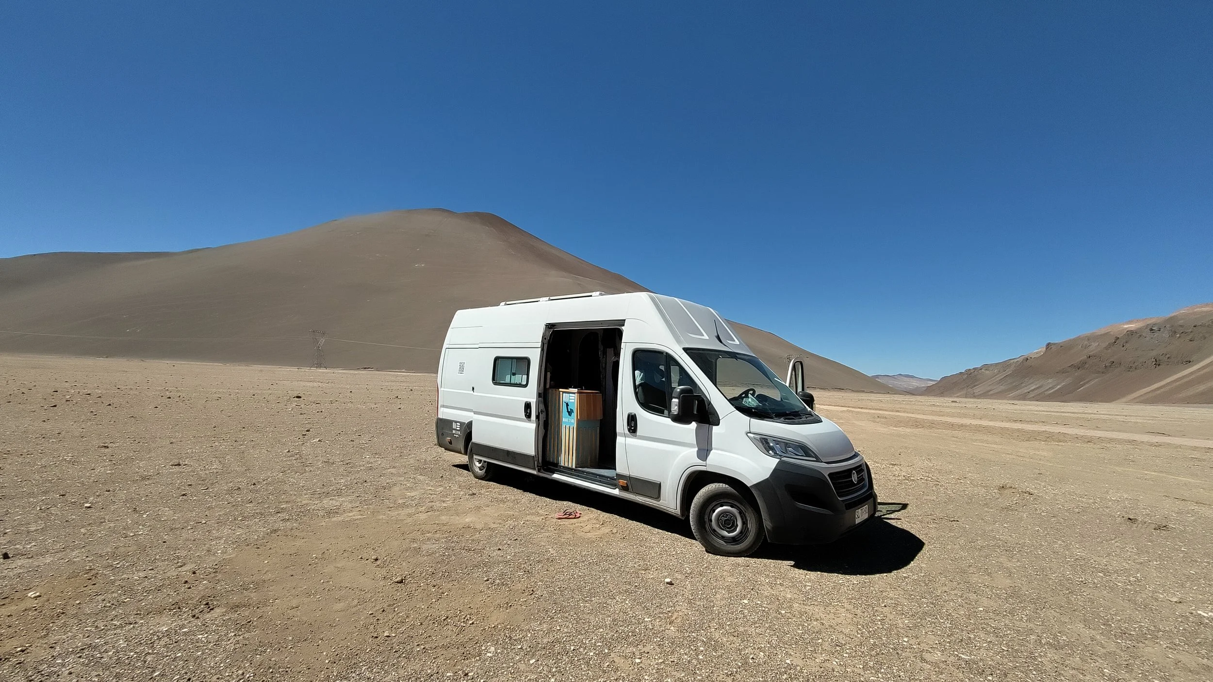 Ein weißer Van steht auf einer trockenen, sandigen Landschaft vor einer Bergkette unter klarem blauen Himmel.