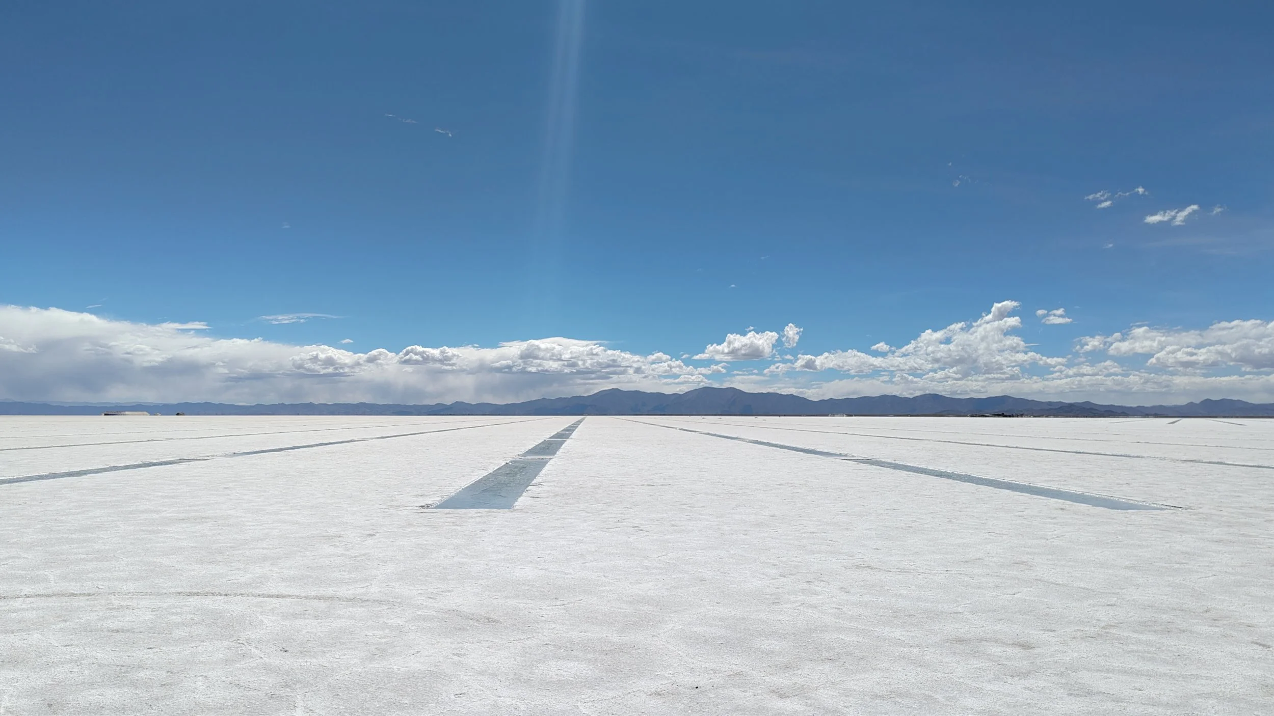 Weißer Salzsee mit Schienen und Bergen im Hintergrund, blauer Himmel mit wenigen Wolken