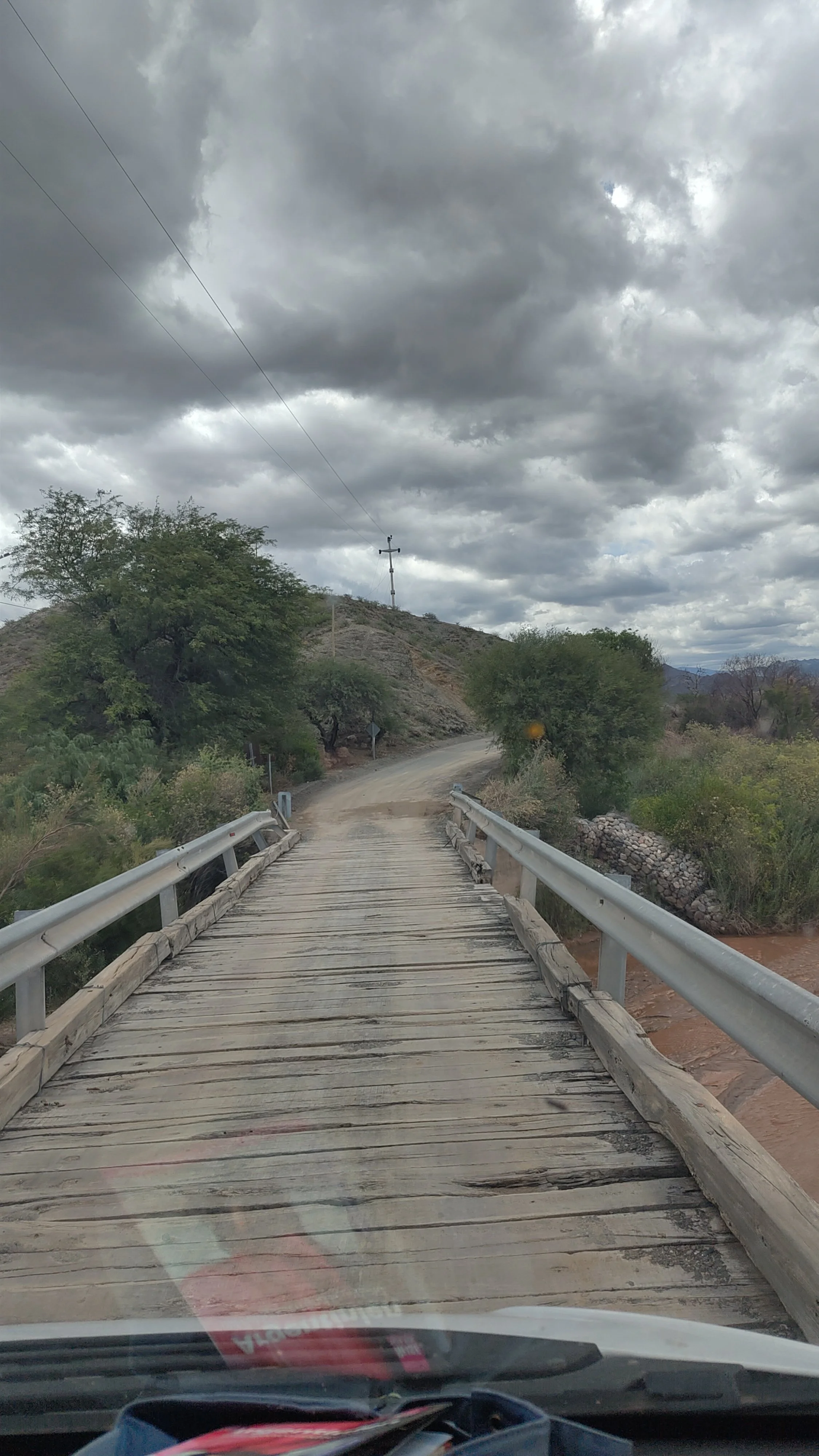 Ein Holzbodenbrücke führt eine Landstraße über einen kleinen Fluss, mit Bäumen und bewölktem Himmel im Hintergrund.