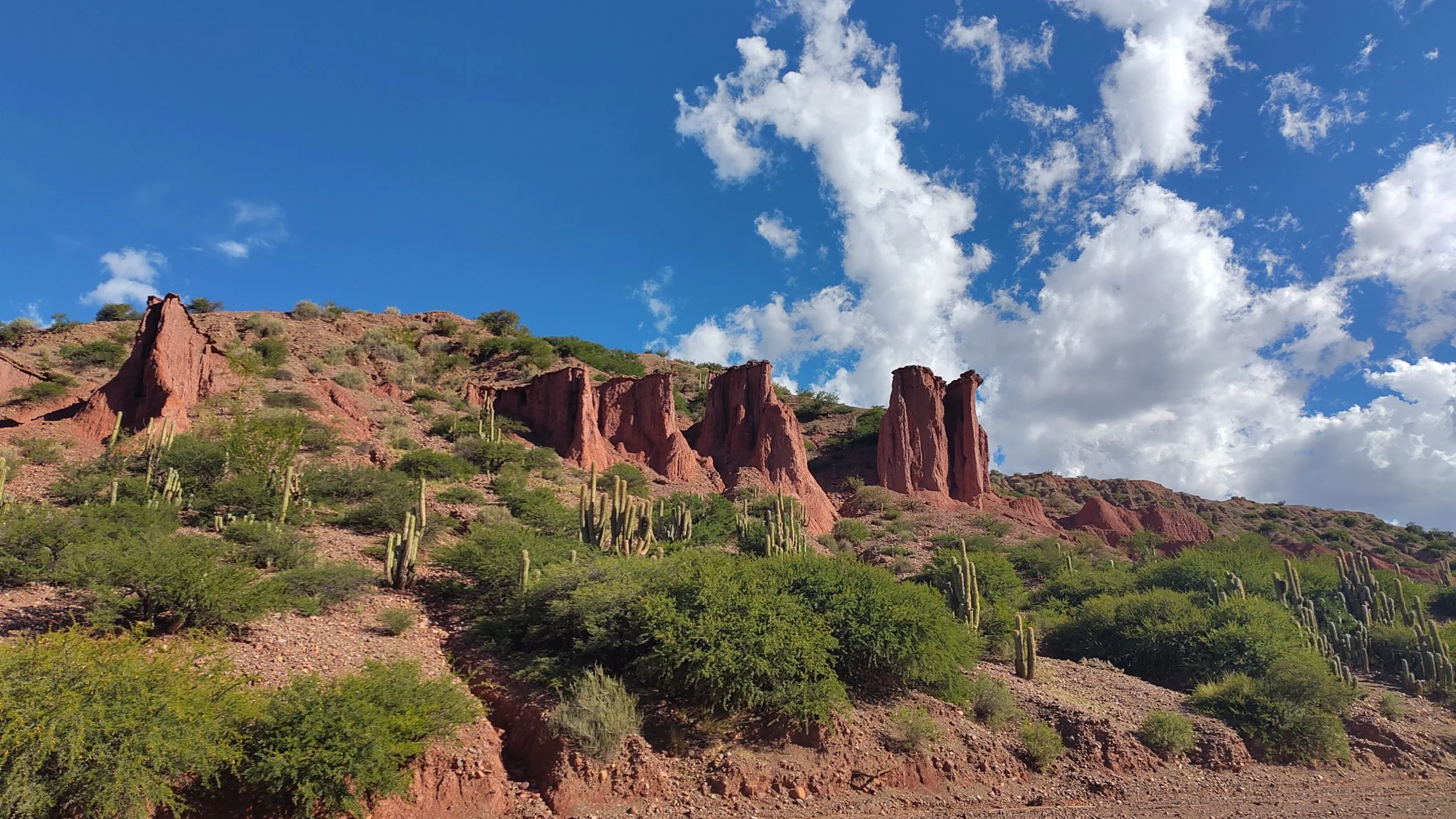 Rote Felsformationen auf einem Berg mit grüner Vegetation und Kakteen, darunter ein blauer Himmel mit weißen Wolken.