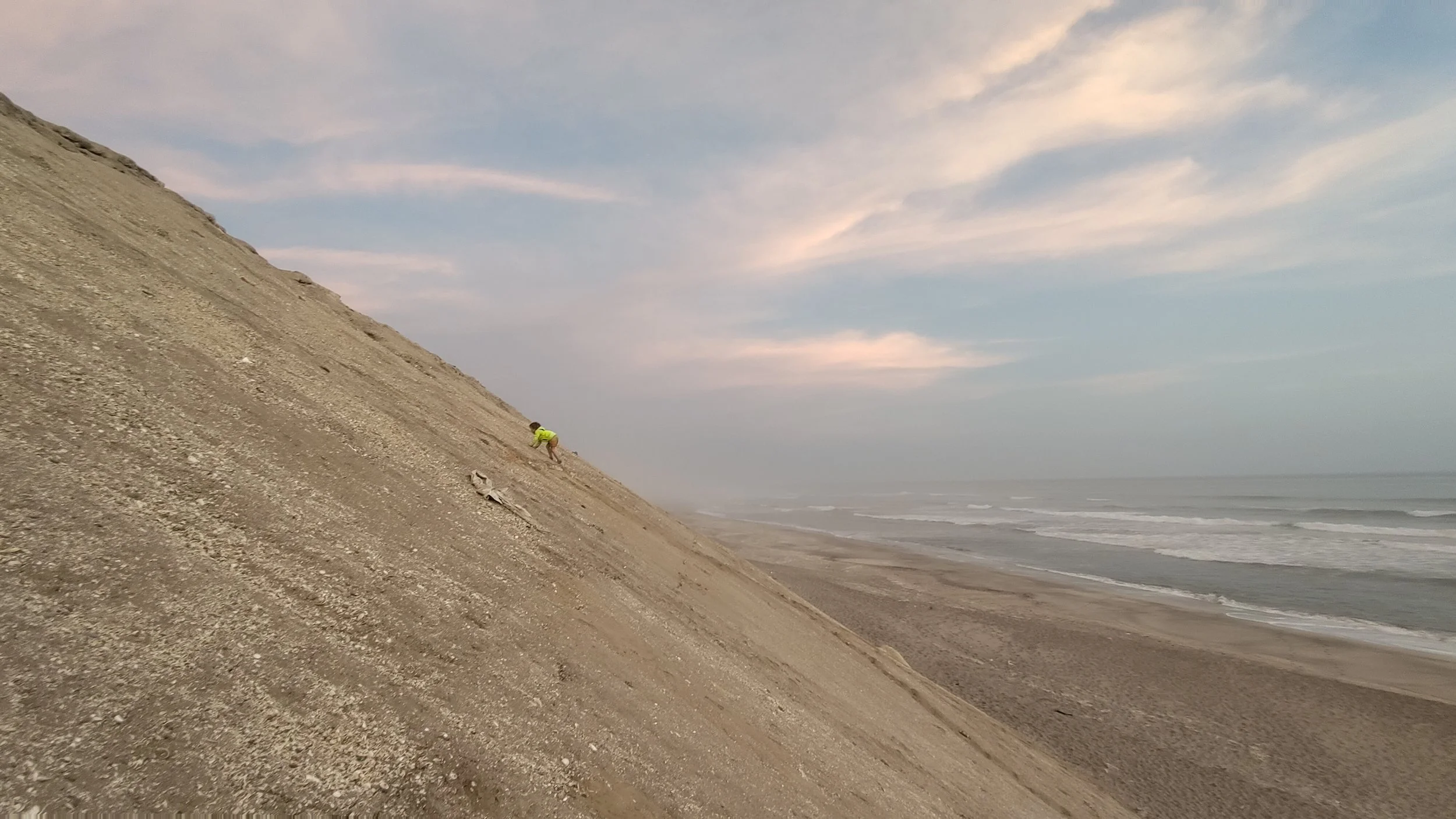 Ein Mensch klettert eine steile, sandige Düne an einem Strand nach oben, mit Blick auf das Meer und den Himmel im Hintergrund.