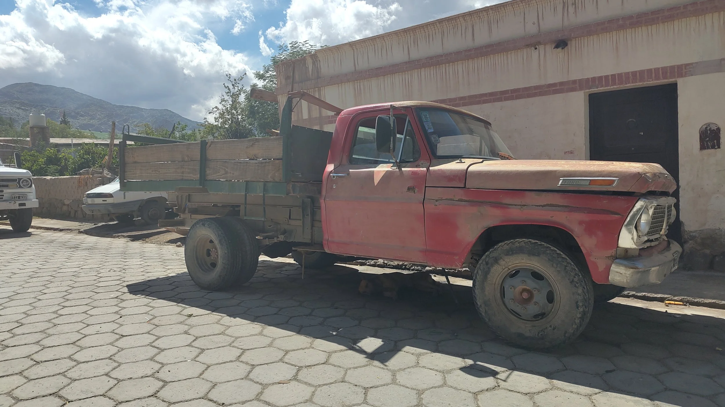 Ein alter, roter Pickup-Truck mit beschädigter Motorhaube auf einer gepflasterten Straße, im Hintergrund Berge und eine weiße Wand