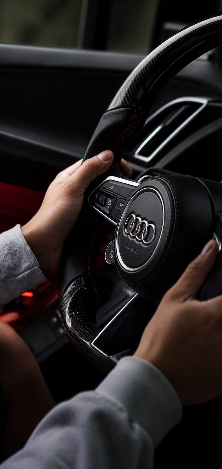 Close-up view of a person holding the steering wheel of an Audi sports car, featuring a carbon fiber grip and buttons on the center.”}