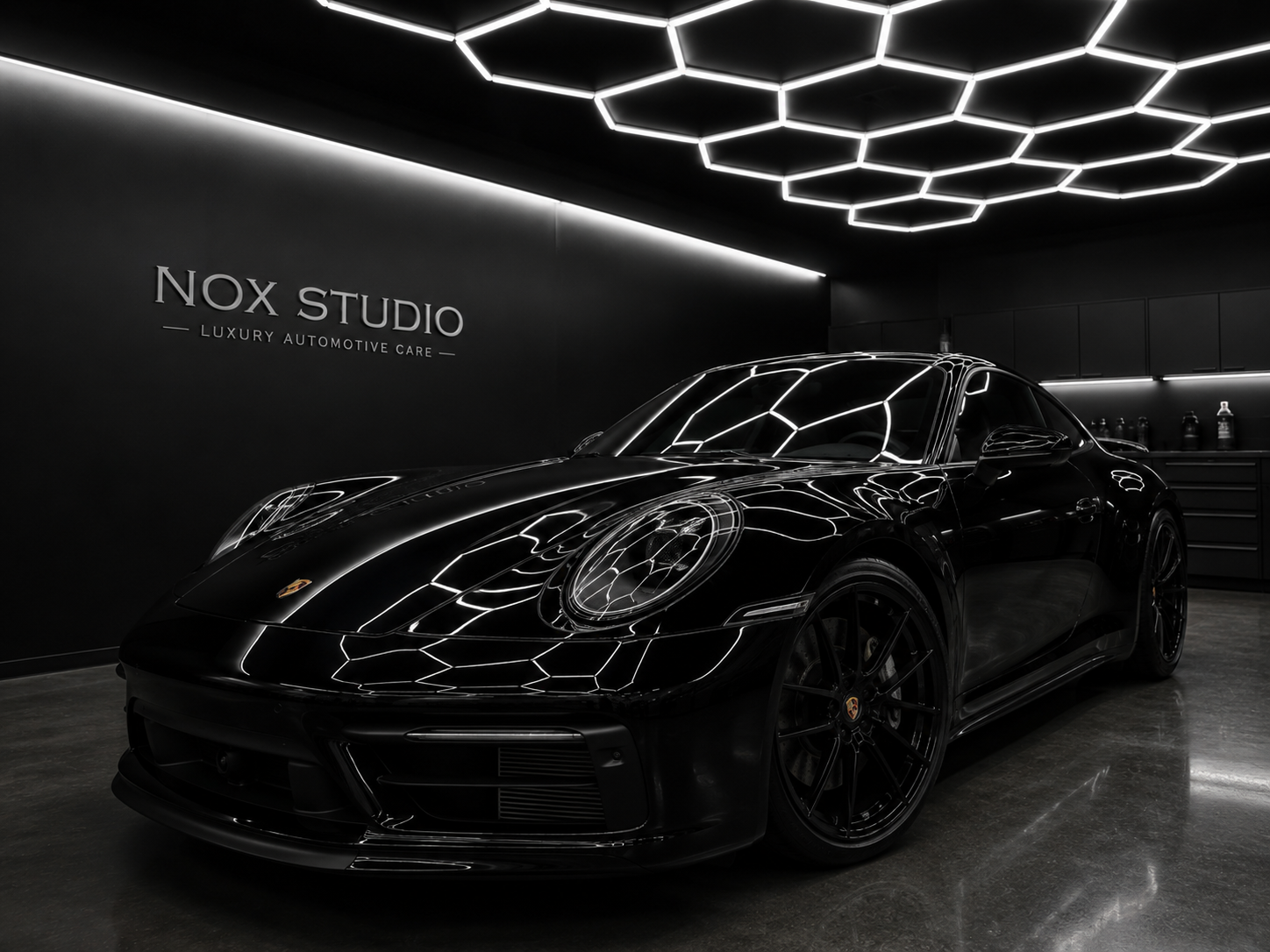 A black luxury sports car displayed in a modern automotive care studio with black walls and hexagonal ceiling lights reflecting on its surface.