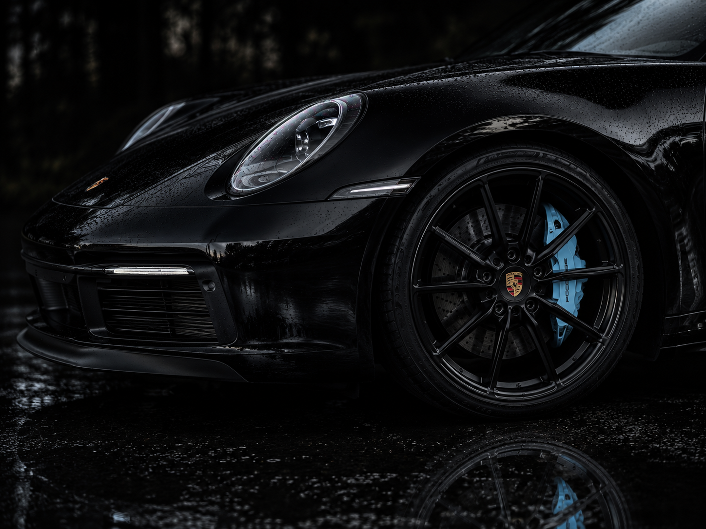 Close-up of a black Porsche sports car with water droplets on its surface, focusing on the front left side, wheel and headlight, on a wet road at night.