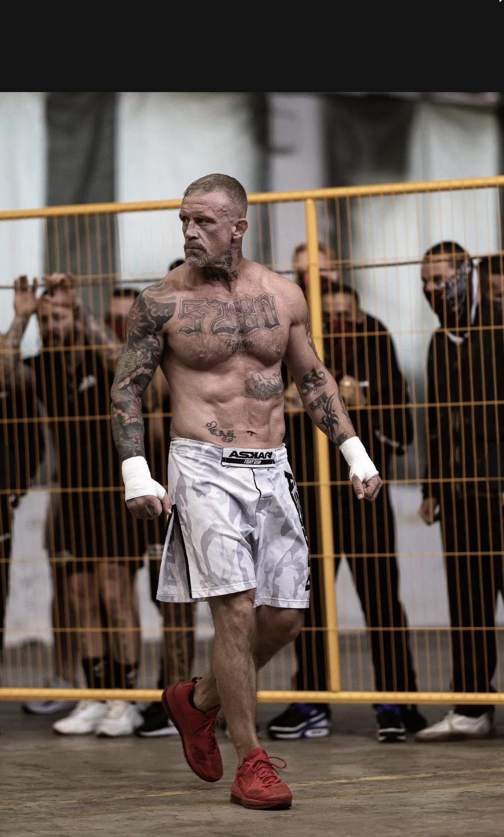 A tattooed male fighter walking in a gym or competition area, wearing white shorts, red shoes, and white hand wraps, with a yellow metal fence in the background and spectators behind it.