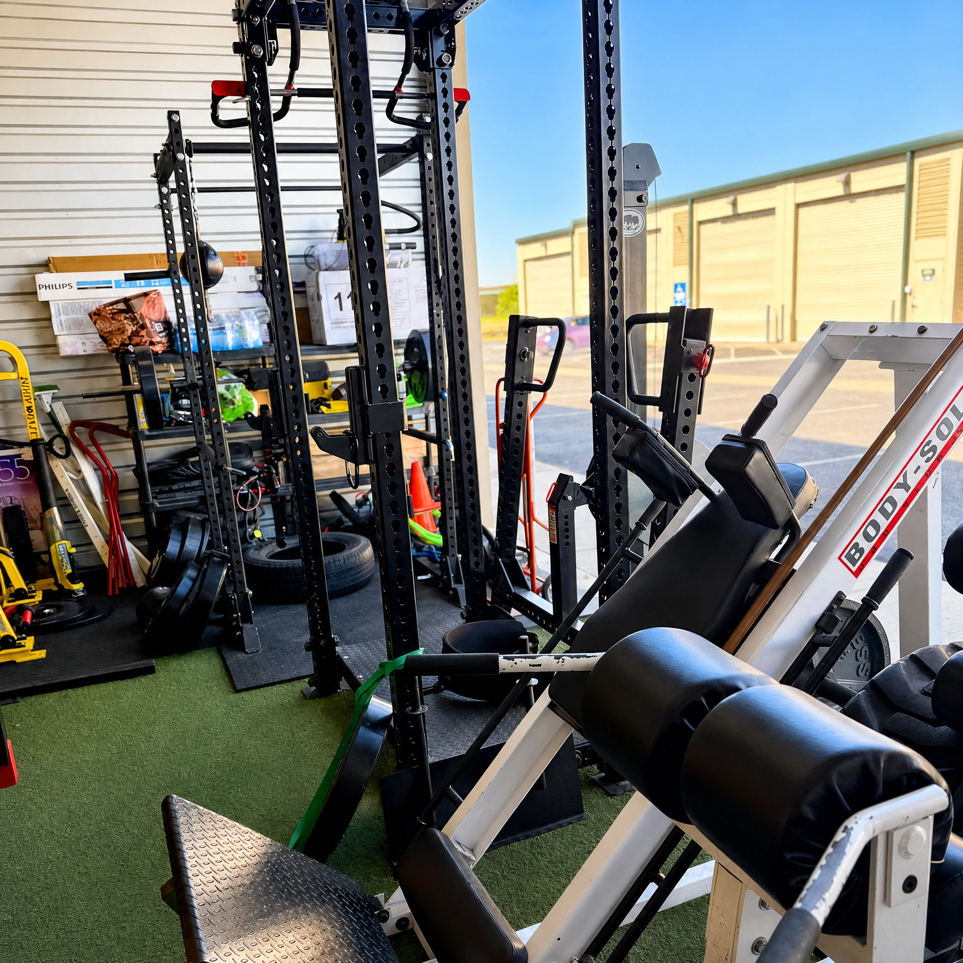 Outdoor gym equipment including weight machines, a bench, and exercise accessories on artificial turf, with a tan building and clear blue sky in the background.