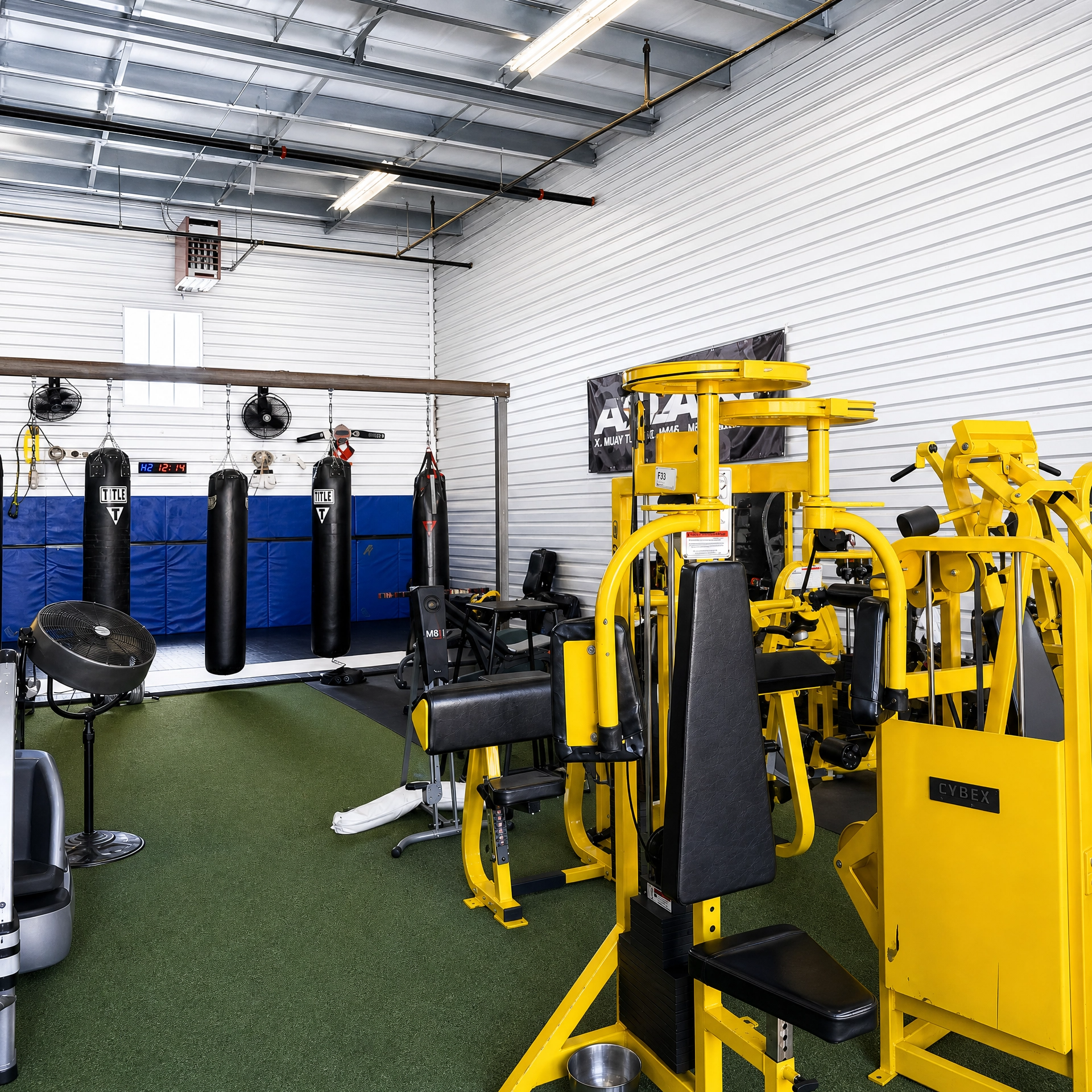 Inside a gym with workout equipment, including yellow exercise machines, punching bags, a fan, and a wall with black gloves and a digital clock.