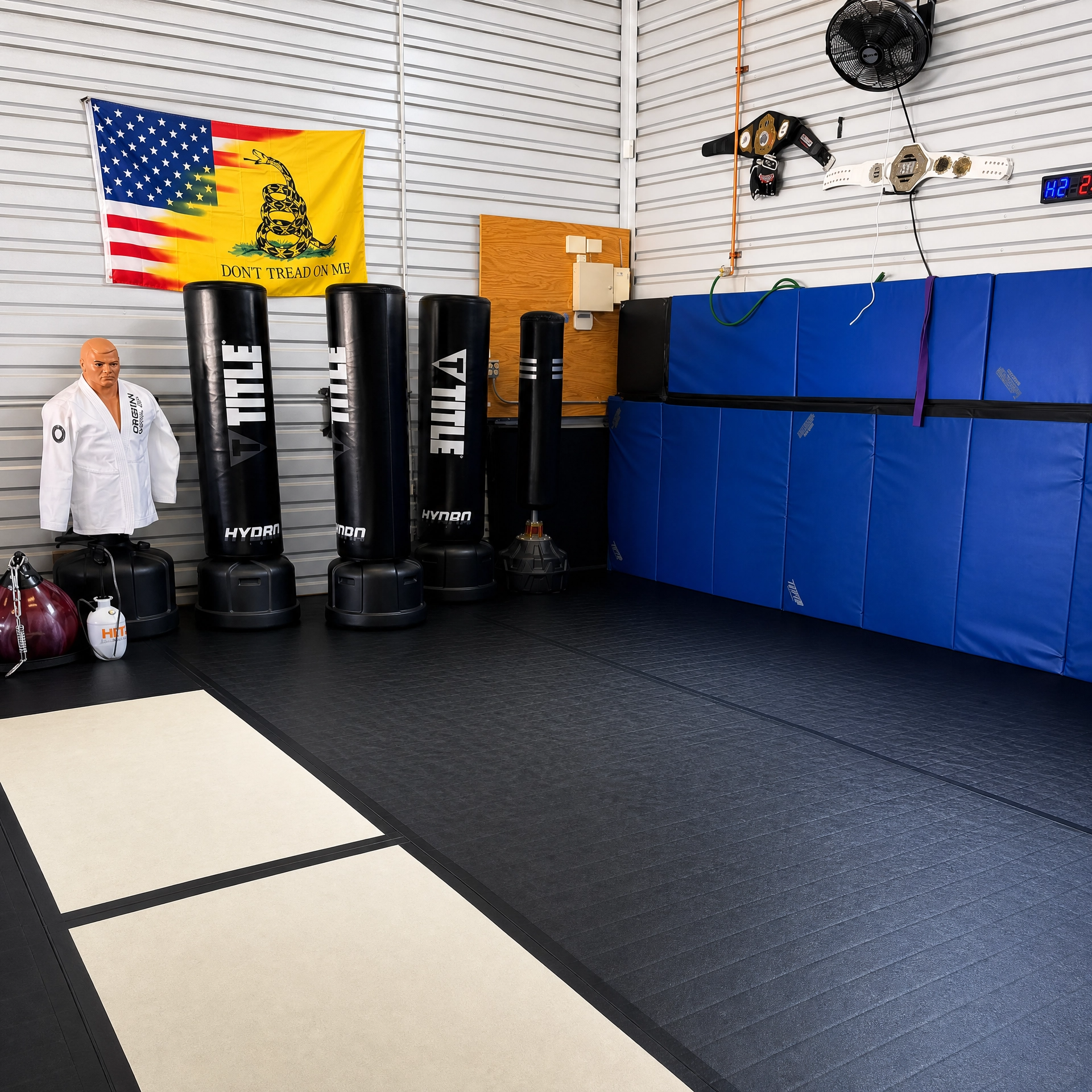 Martial arts gym with a white gi mannequin standing on black flooring, three black punching bags with white labels, a blue padded wall with purple and green belts, a yellow flag with red and blue elements, gloves, belts, a scoreboard, and a ceiling f