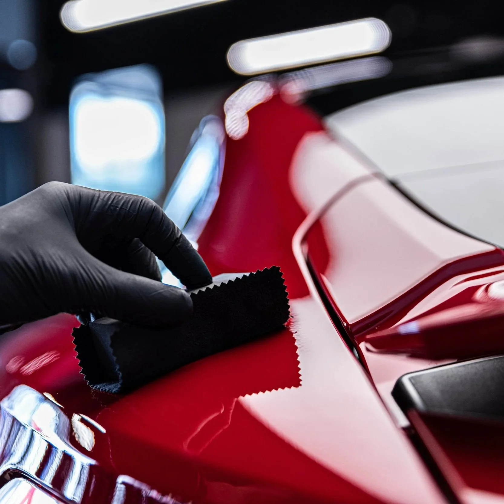 A person using a ceramic coating applicator to apply coating on red exterior of a car, with some reflections visible on the car's shiny surface.