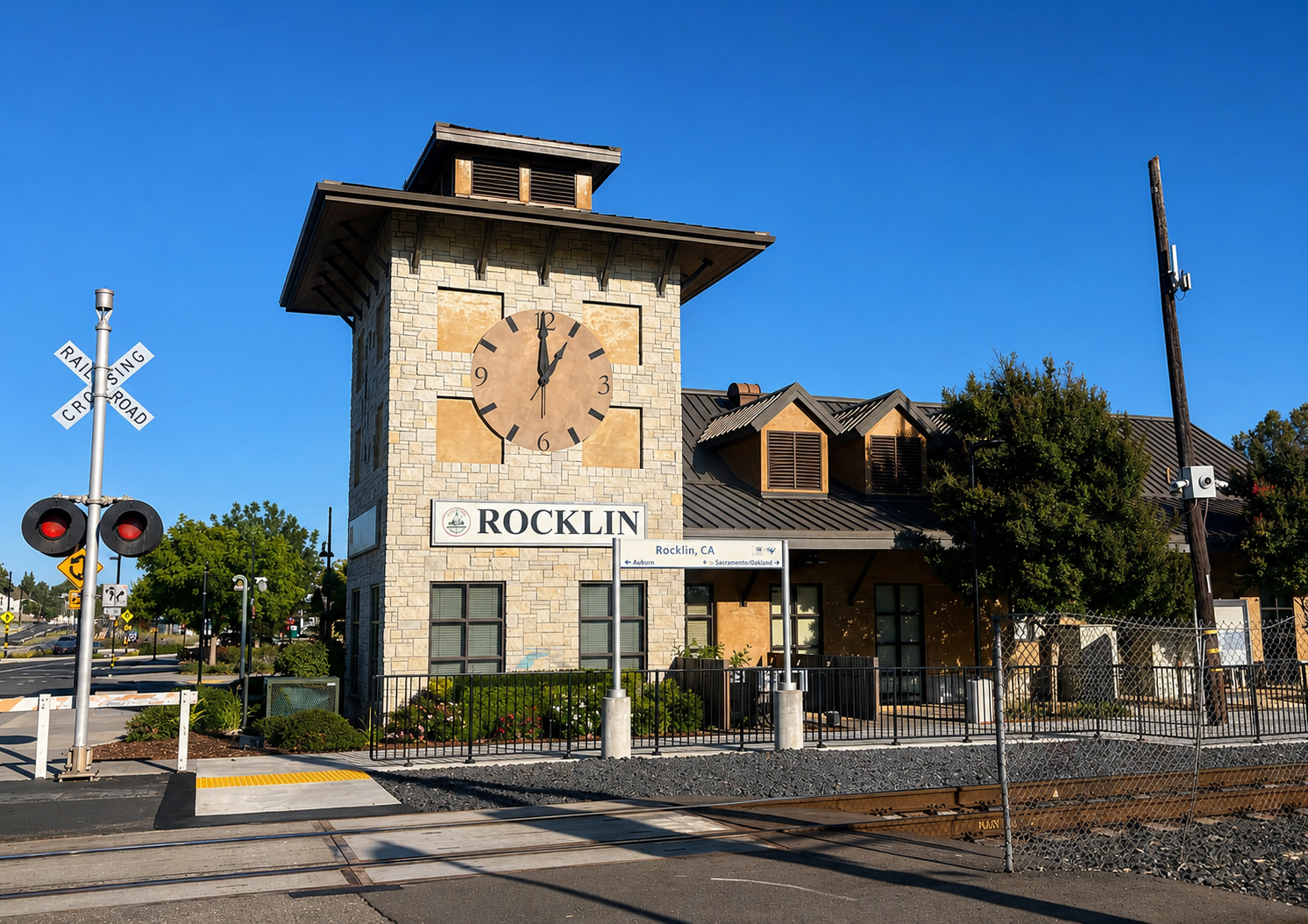 A train station with a brick clock tower showing the time at 12:00, a railroad crossing with signals, and a sign reading 'Rocklin' in Rocklin, California, against a clear blue sky.