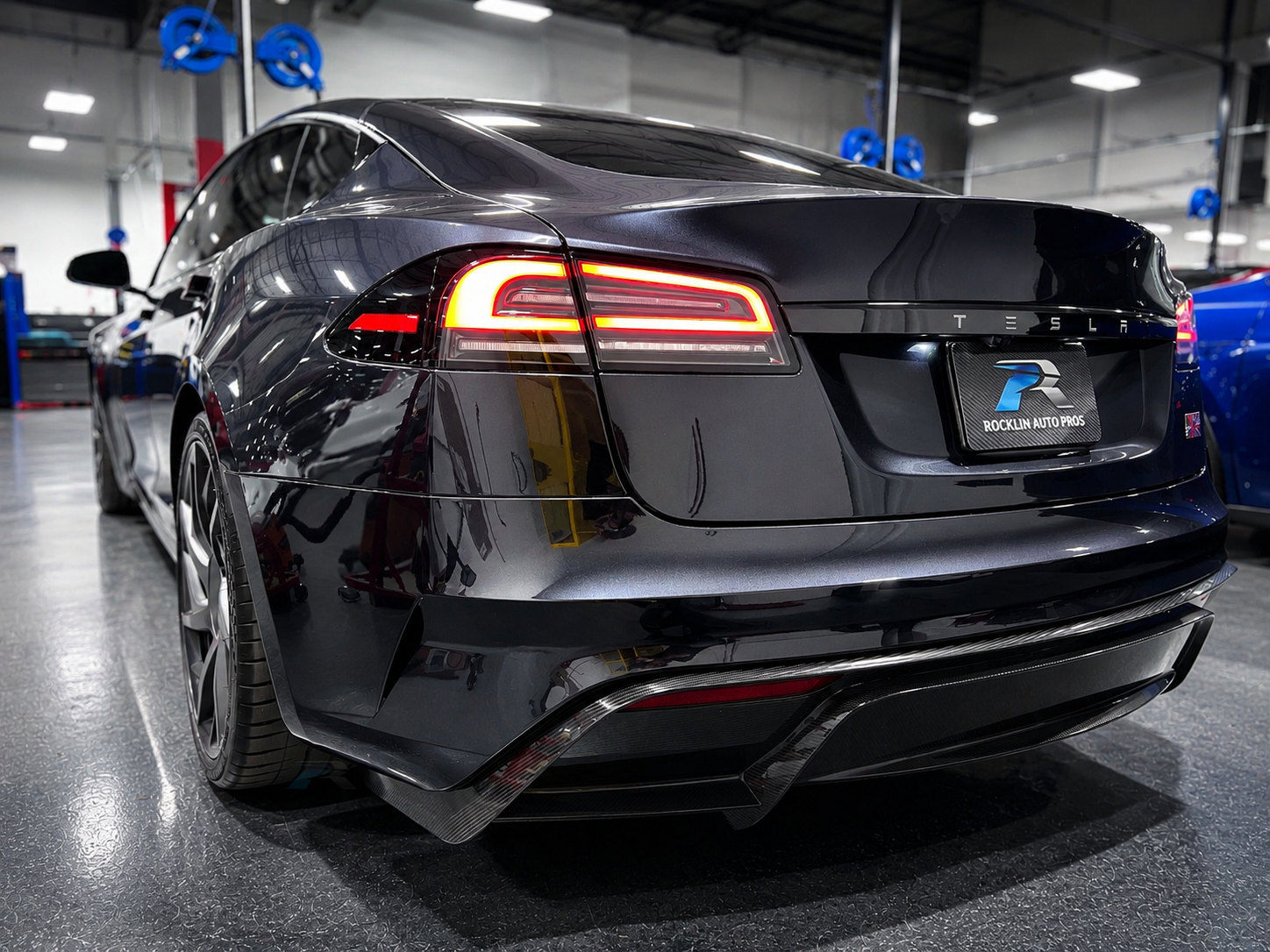 A black Tesla Model S parked in a showroom or garage, with a Rocklin Auto Pros license plate frame, and reflections of overhead lights on the car's surface.