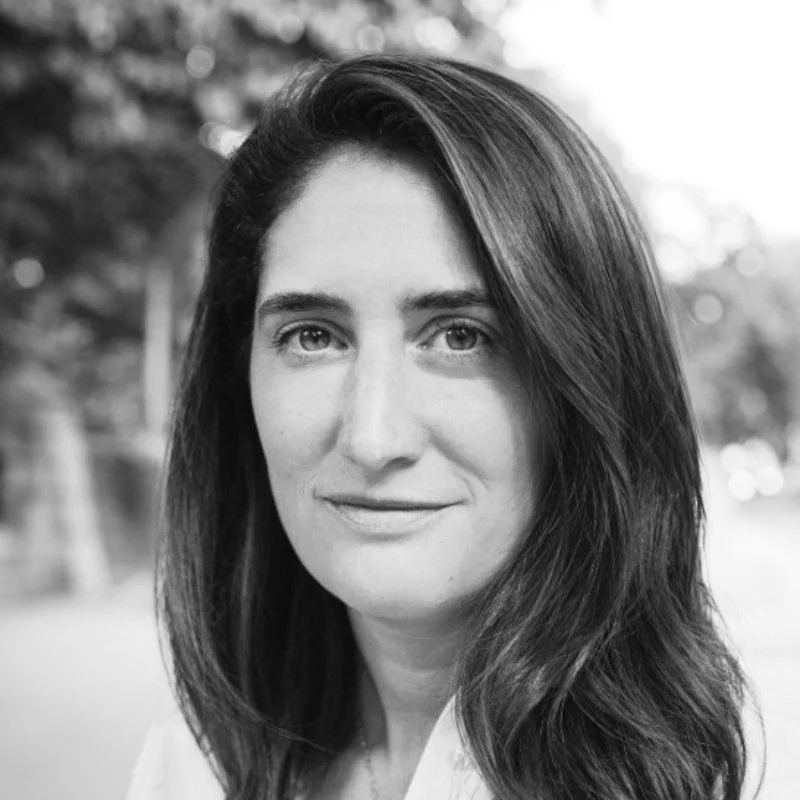 Black and white portrait of Alex, a woman with long dark hair, looking at the camera with a slight smile, outdoors with blurred trees in the background.
