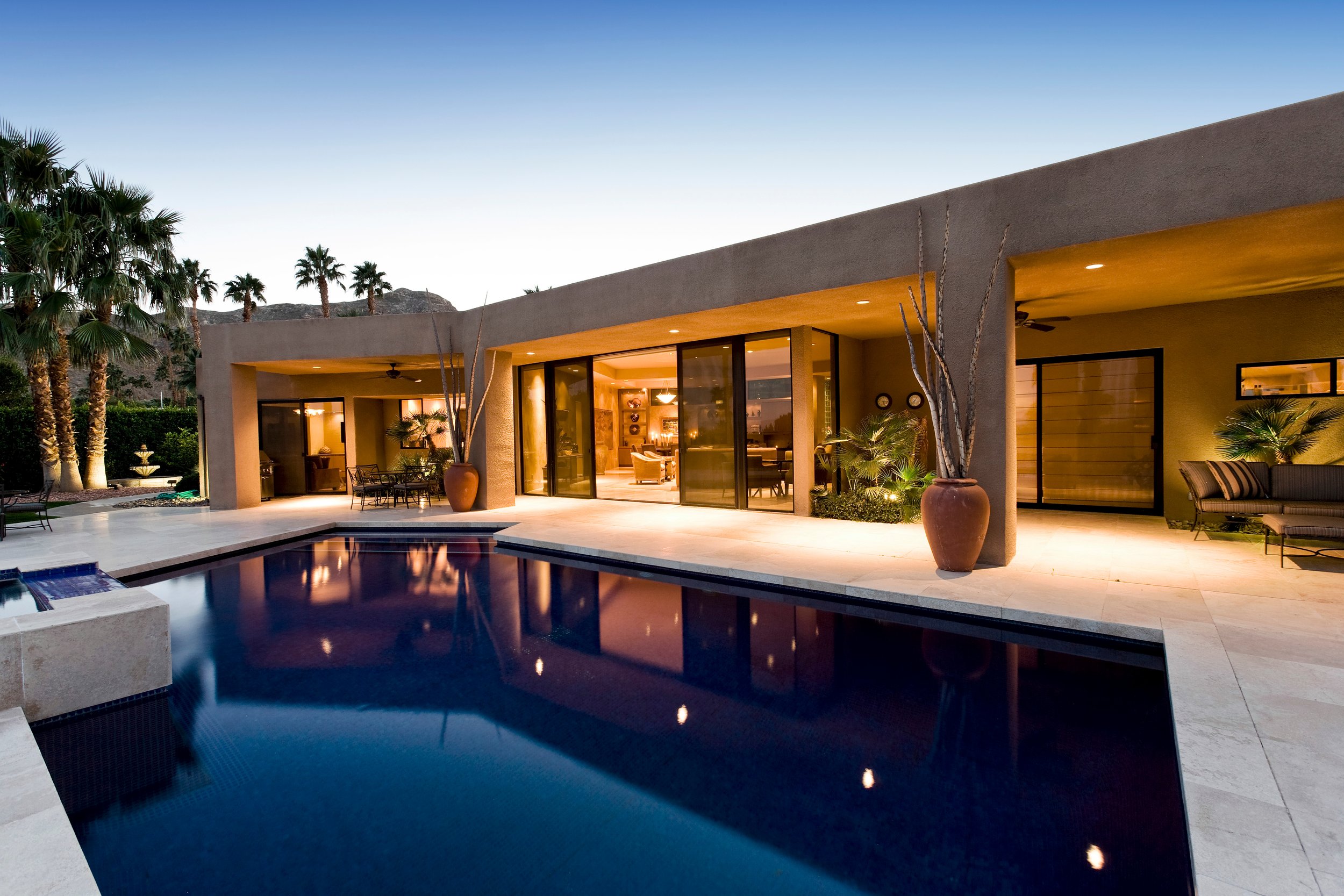 A modern house with large glass sliding doors, a swimming pool, outdoor seating, potted plants, and palm trees in the backyard at dusk.