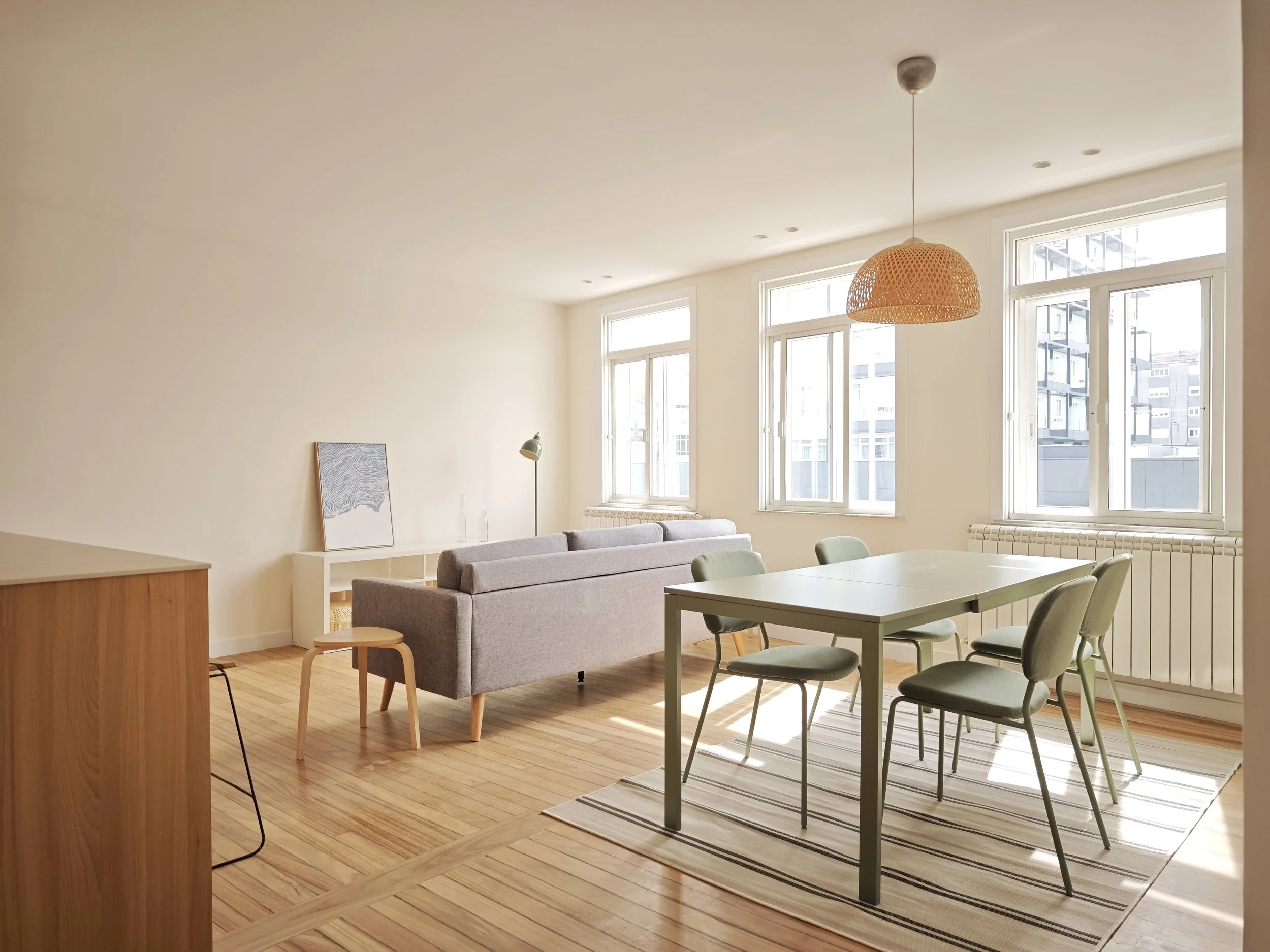 Sala de estar luminosa con ventanas grandes, sofá gris, mesa de comedor, sillas verdes, lámpara de tech o de mimbre, piso de madera y cuadro en la pared.