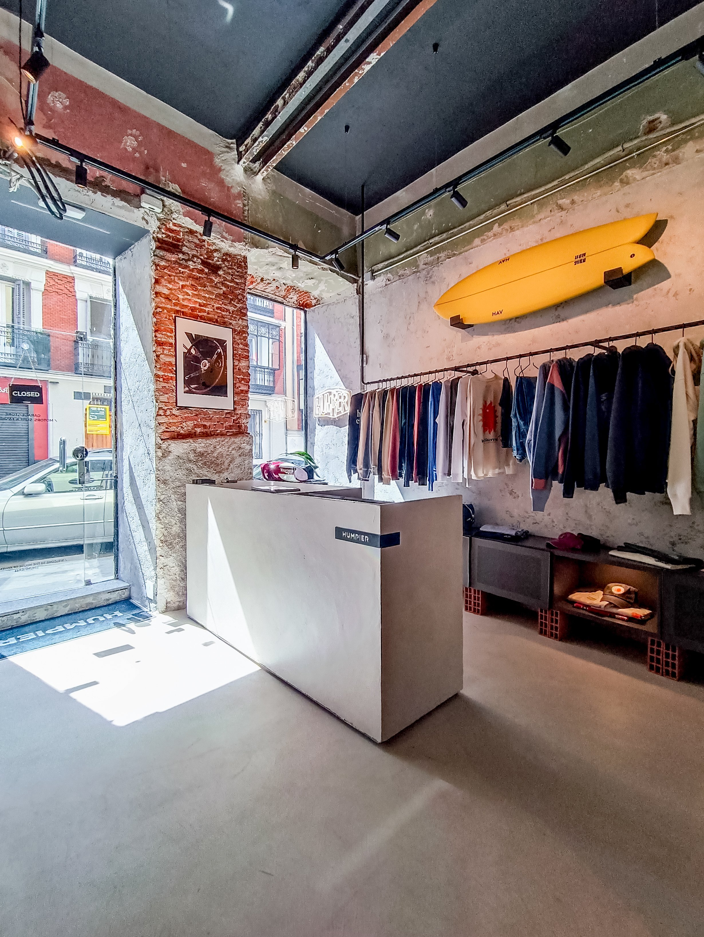 Interior de una tienda con pared de ladrillos expuestos y ropa colgada, exhibidor de surf en la pared, ventana desde donde se observa un coche estacionado afuera.