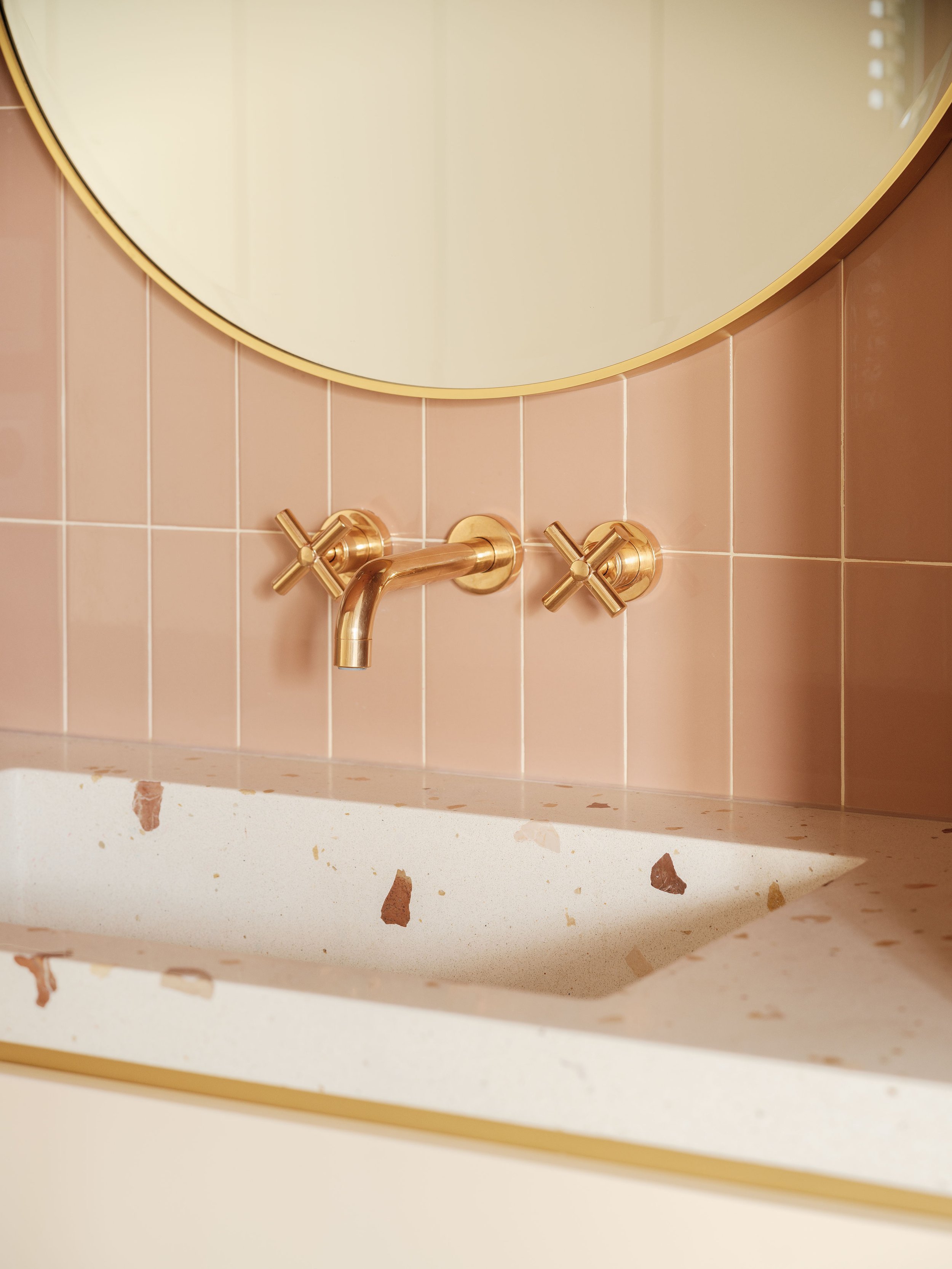 Espejo redondo con marco dorado, dos llaves de grifo doradas y una tubería en una pared de azulejos rosa, y parte de un lavabo con encimera de piedra con manchas.