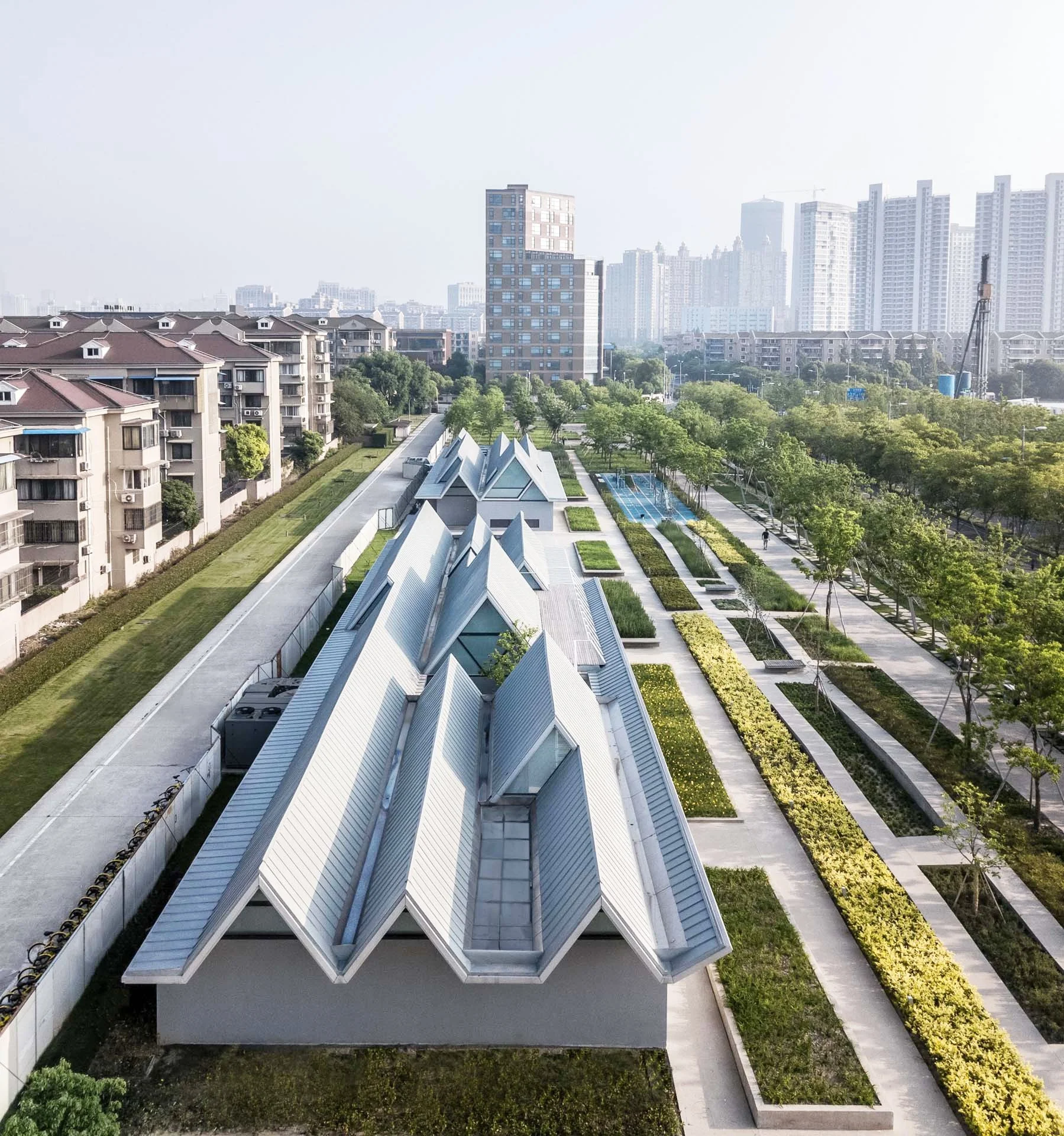 Edificio moderno con estructuras de techos en forma de zigzag, rodeado de áreas verdes, caminos peatonales y árboles, en un paisaje urbano con edificios altos al fondo.