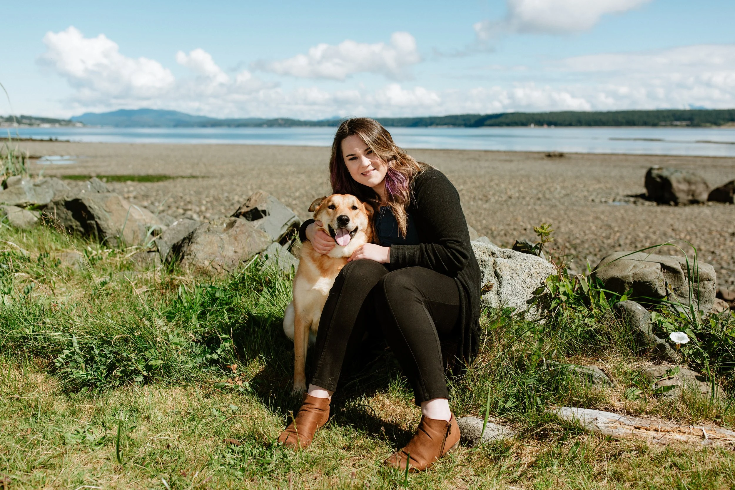 A woman sitting on rocks by a lake with her dog, both smiling, with a scenic view of water, distant mountains, and partly cloudy sky in the background.