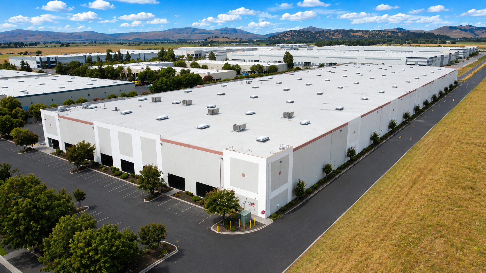 Aerial view of a large industrial warehouse with a white roof, surrounded by parking lots and trees, with mountains in the background.
