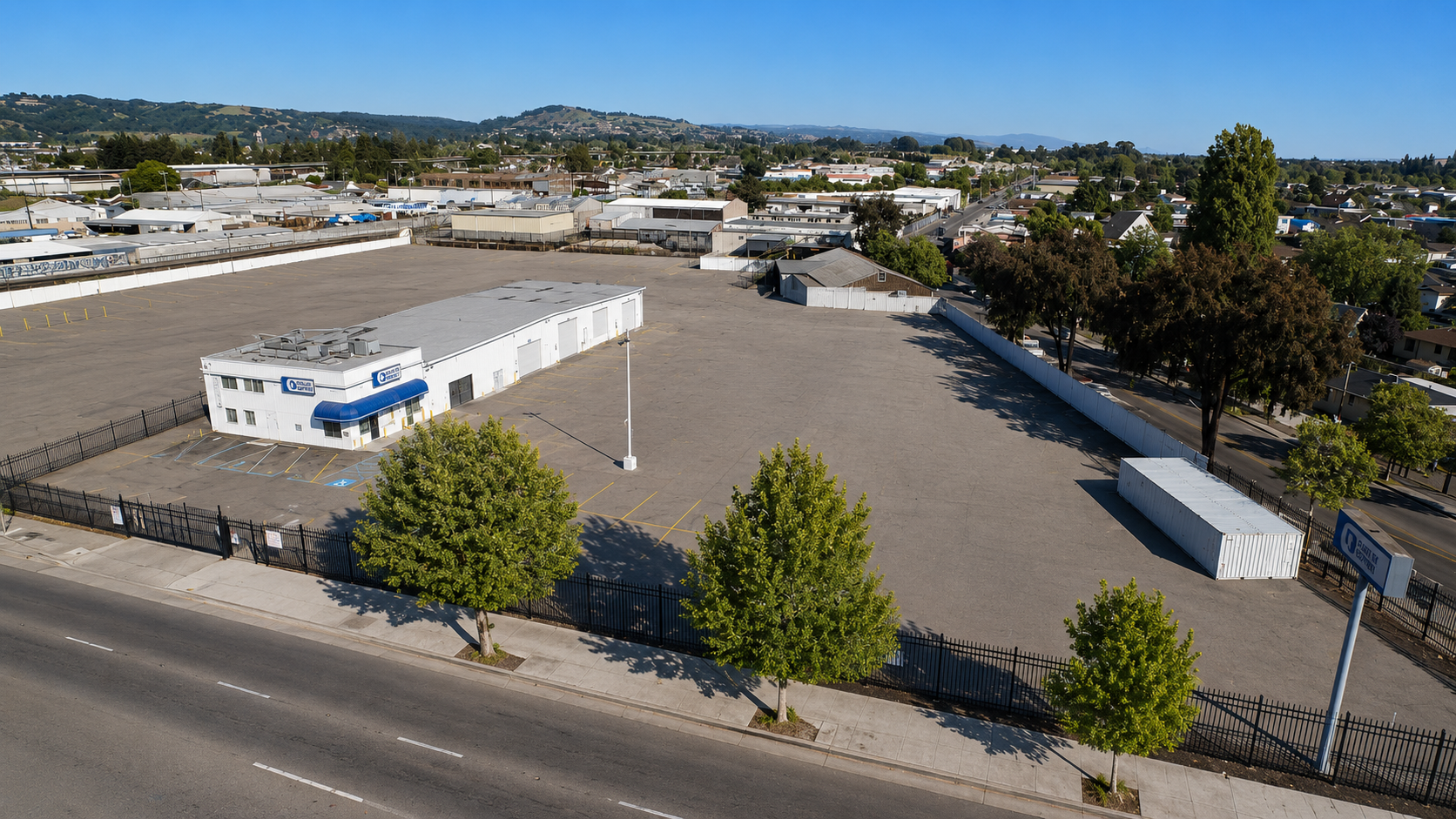 Empty commercial parking lot with a small building, surrounded by trees and fencing, with a cityscape and hills in the background.