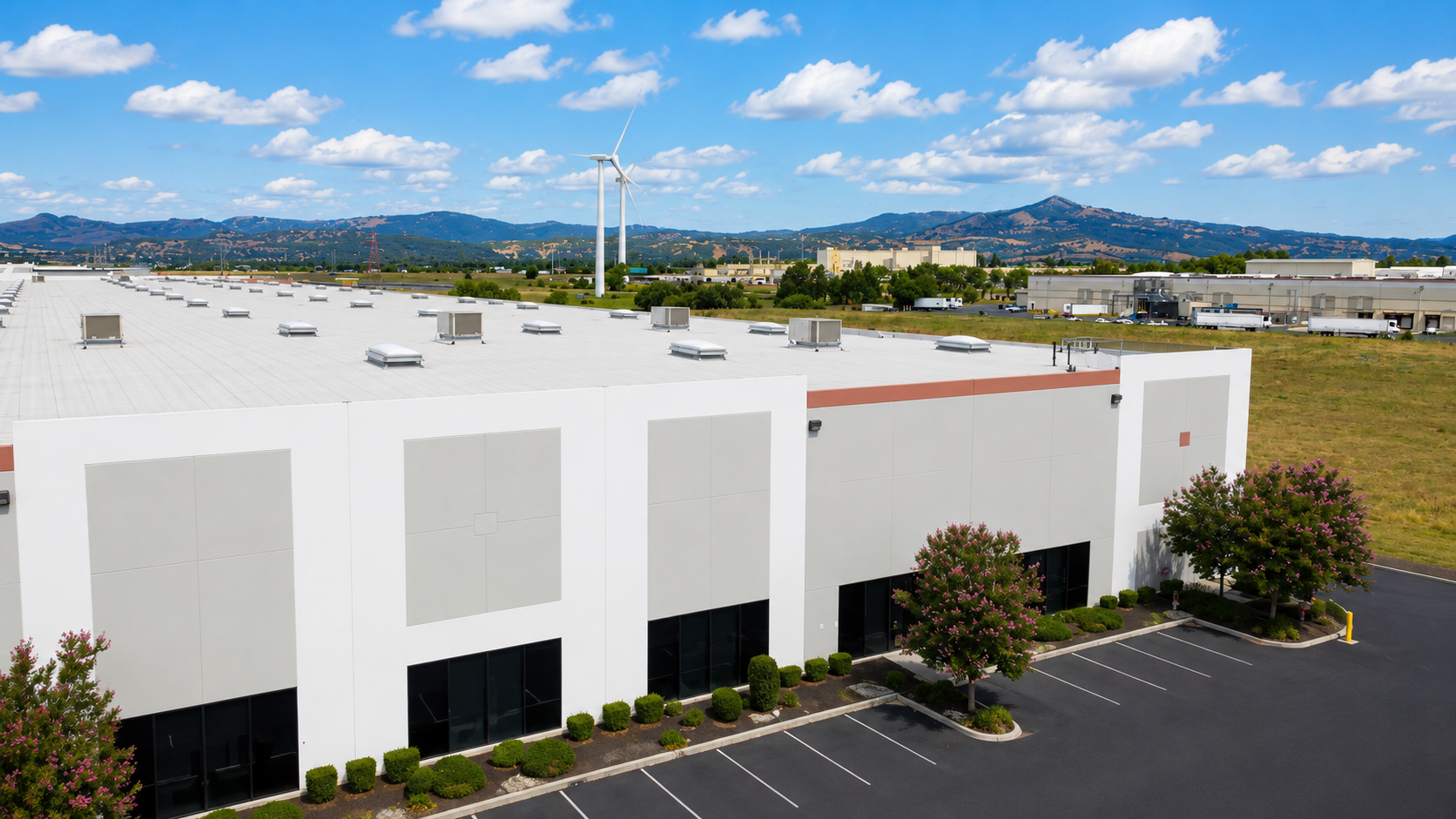 An industrial building with a white roof, surrounded by a parking lot with a few trees and bushes, wind turbines in the background, and mountains under a partly cloudy sky.