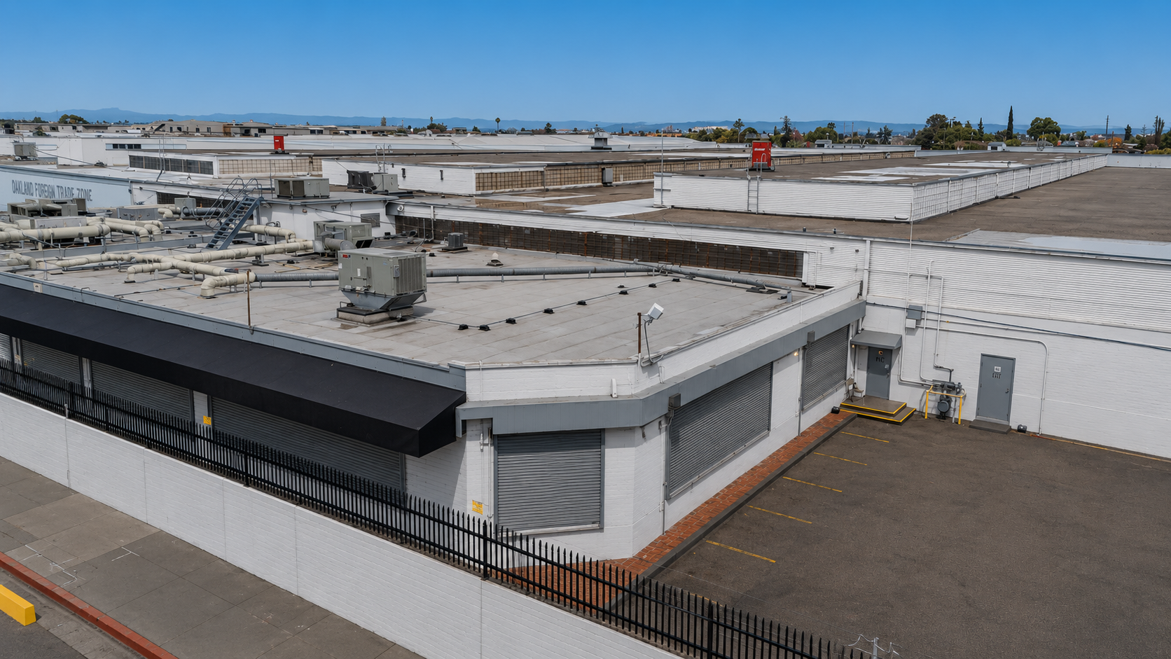 A flat roof of a commercial building with HVAC units, vents, and piping, surrounded by a black fence, with an empty parking lot and a cityscape in the background.