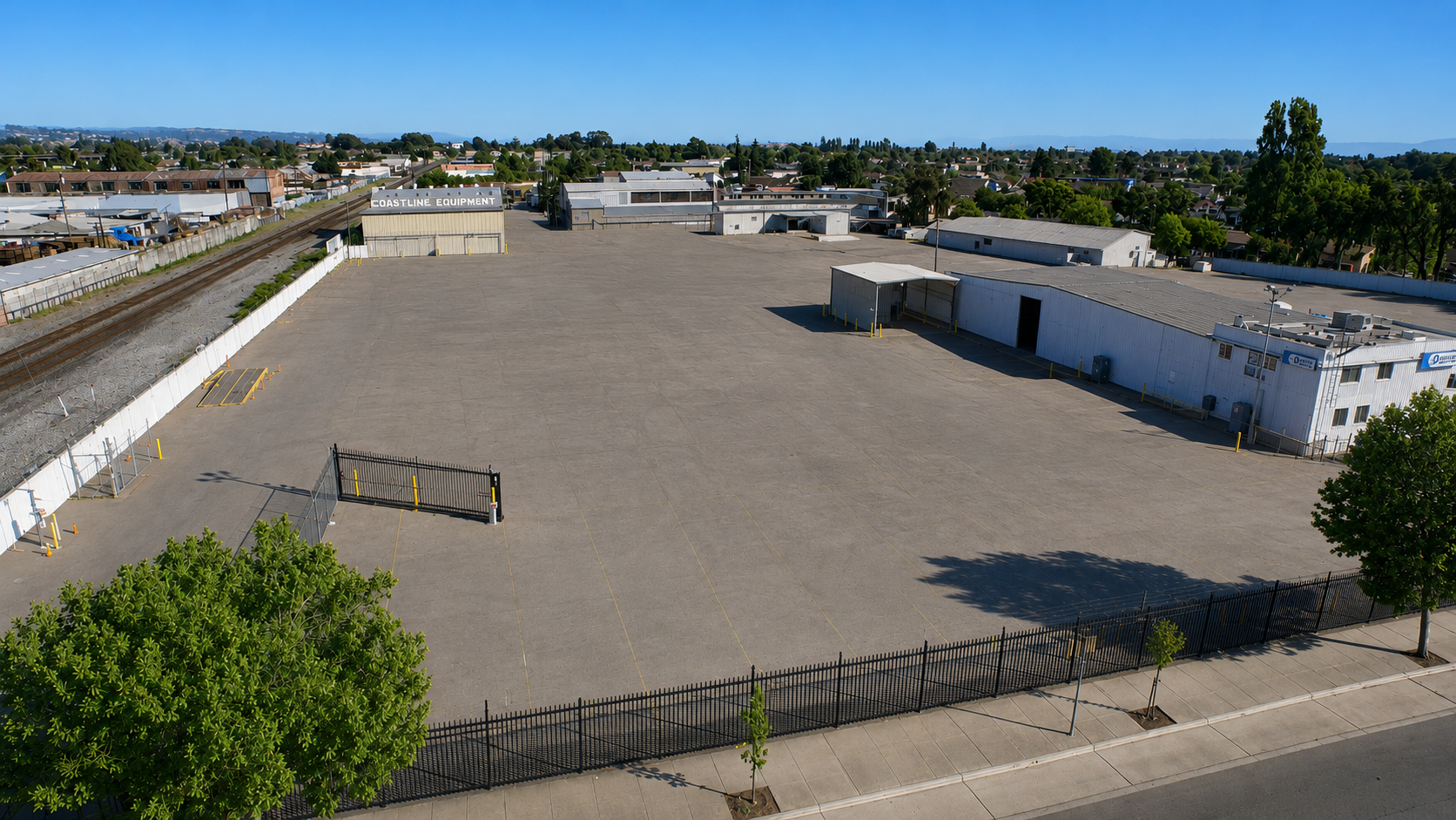Empty parking lot surrounded by a black fence, with buildings and street trees nearby, and train tracks with a sign that says 'Coastline Equipment' in the background.