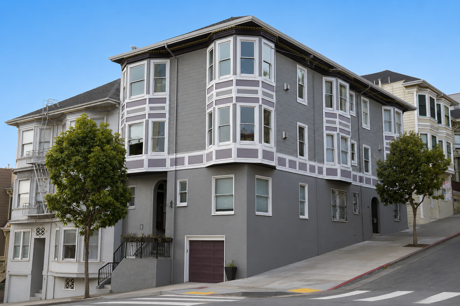 Multi-story residential building with gray and white exterior, bay windows, and trees in front, on a sloped street.