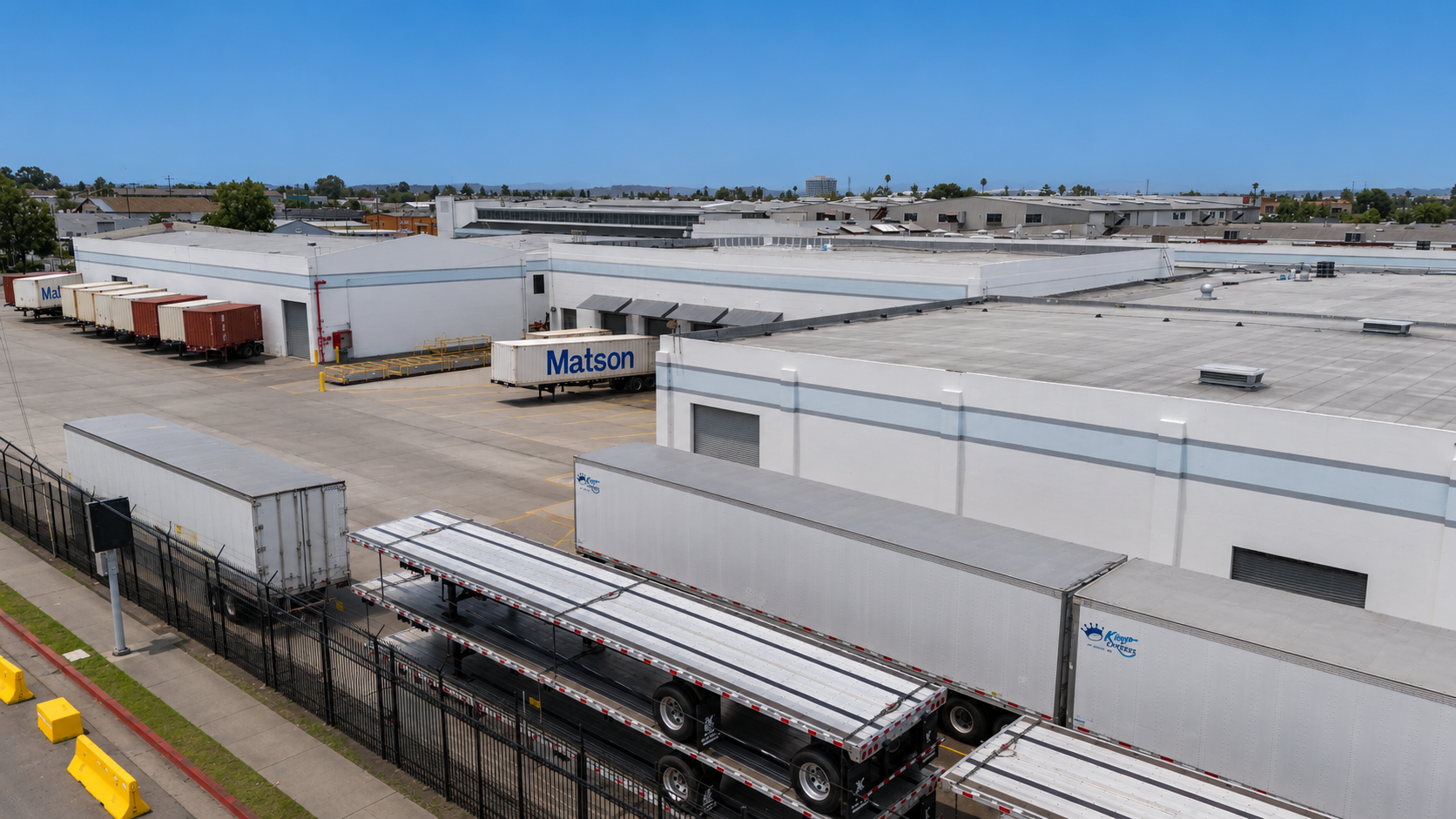 View of a large commercial warehouse with multiple loading dock doors, trucks, and trailers parked outside, including a Matson container, behind a fenced area with yellow concrete barriers on a clear day.