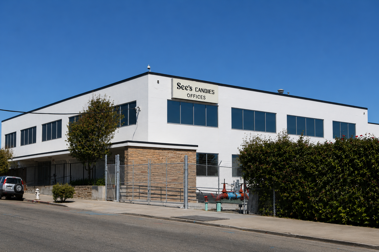 A white, two-story office building with large windows, situated on a corner lot with trees and bushes. A sign on the building reads 'See's Candies Offices'. There is a parked car on the street and a fence surrounding part of the property. The sky is clear and blue.