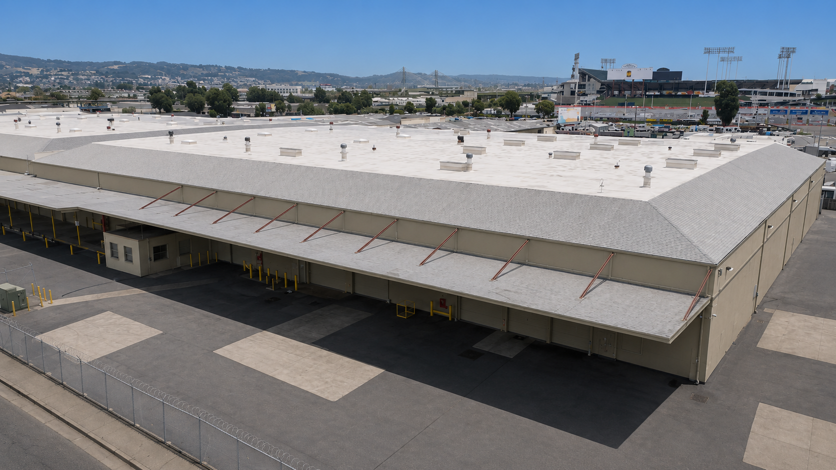 A large industrial building with a flat roof and surrounding paved areas, set against a hilly background.