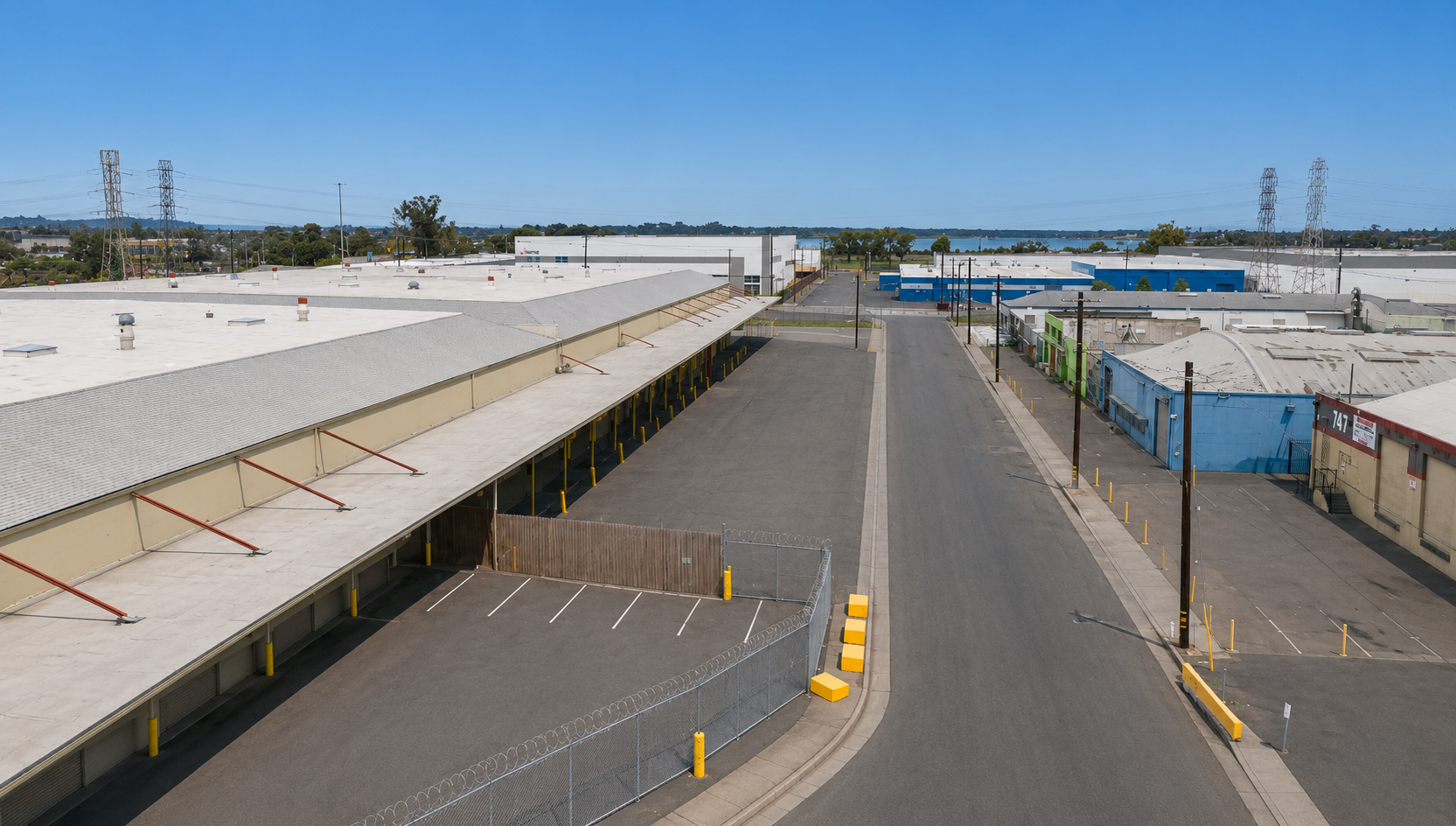 Empty industrial loading dock area with warehouses, parking spaces, and utility poles under a clear blue sky.
