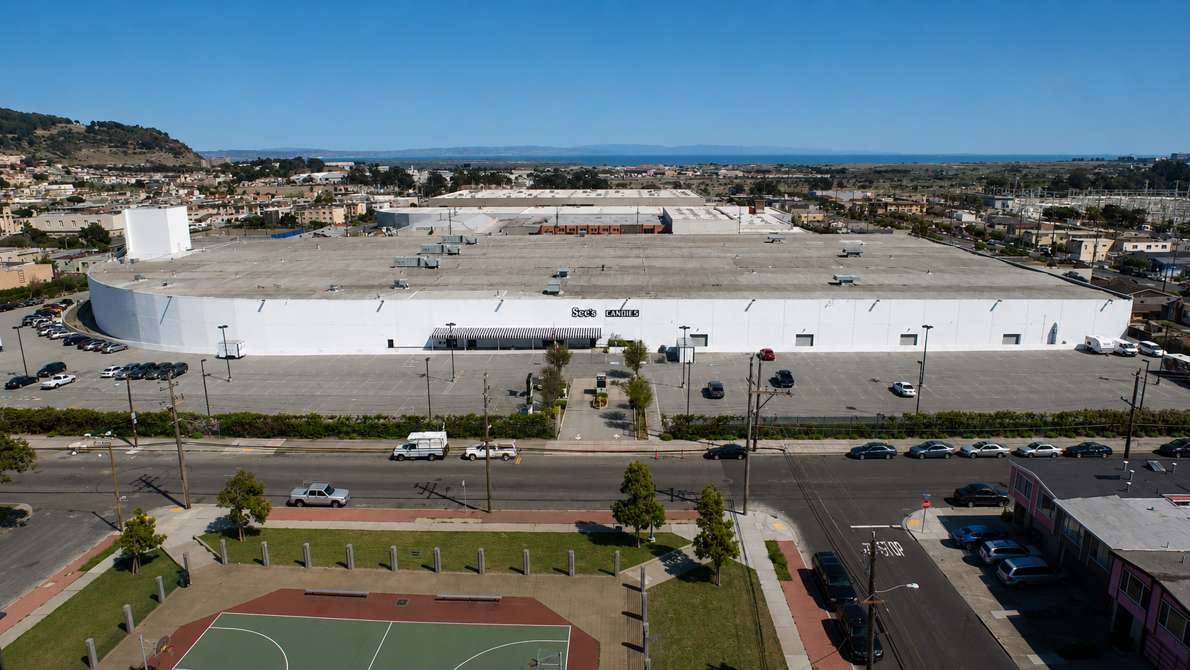 Aerial view of a large white commercial building with parking lot, in a city with houses and distant mountains and water in the background.