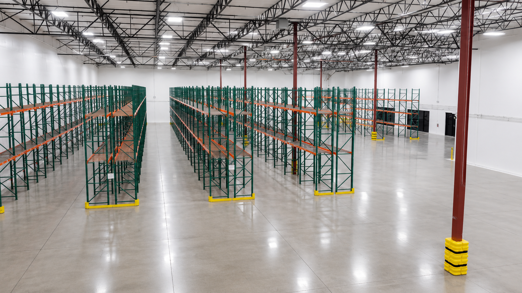 Empty warehouse with multiple green and orange metal storage racks, clean polished concrete floor, white walls, and a high ceiling with exposed black metal beams and bright overhead lighting.