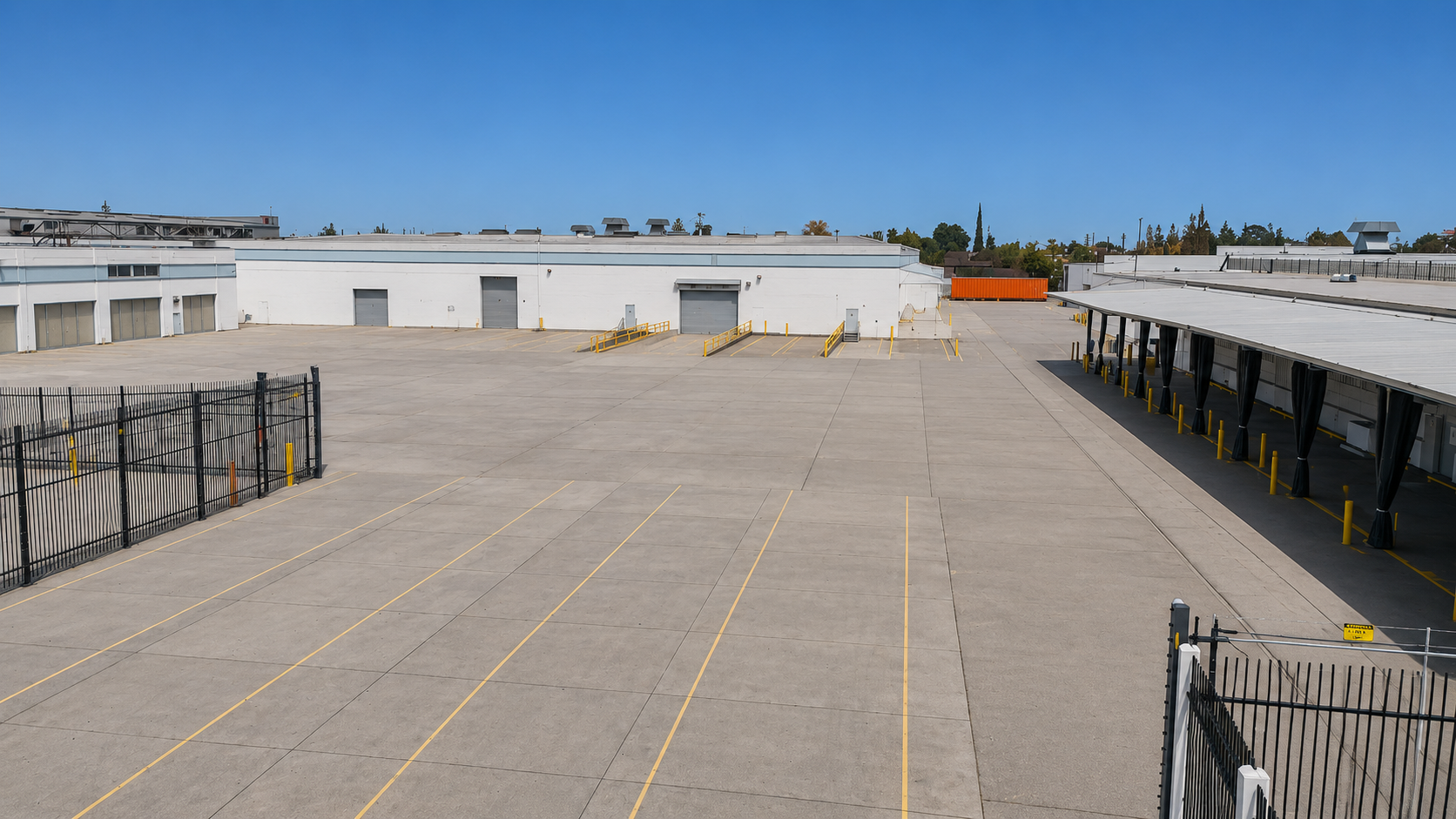 Empty parking lot outside warehouses with a clear blue sky and distant trees.