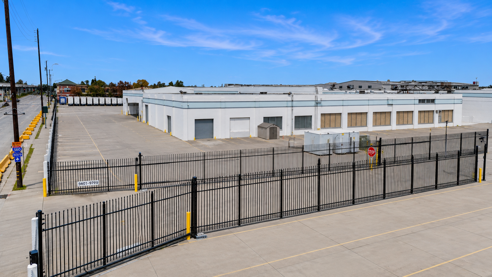Empty parking lot with a white warehouse building, black fence, stop sign, yellow barriers, and a few trucks in the distance under a blue sky.