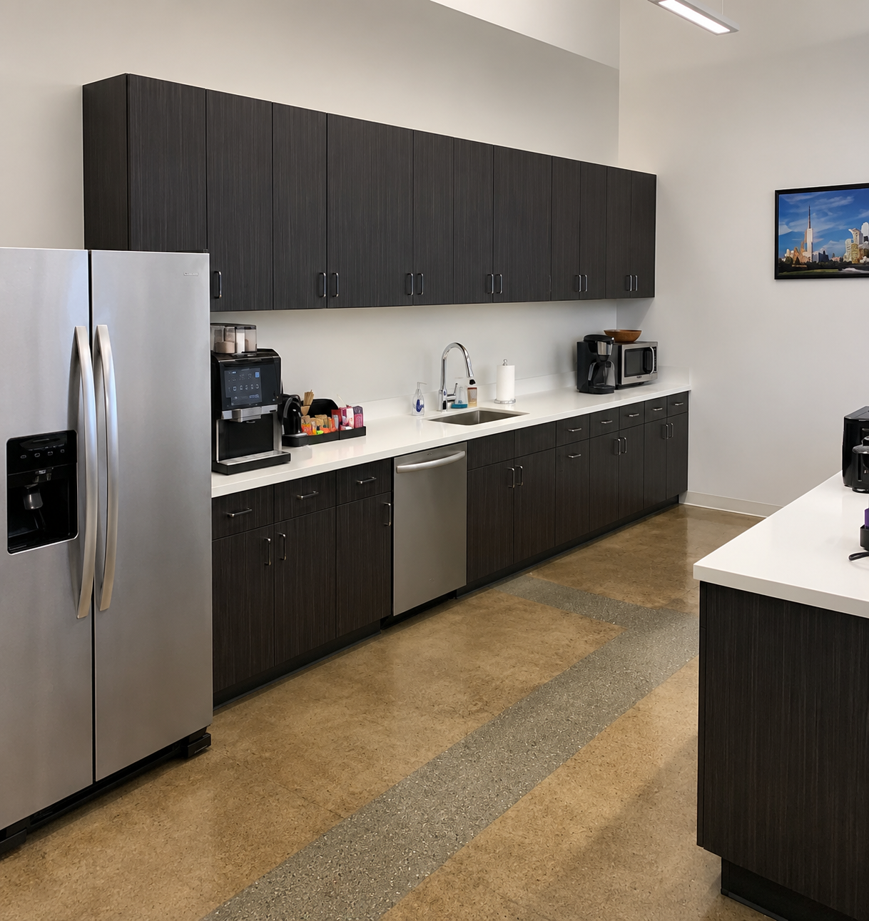 Modern kitchen with dark wooden cabinets, stainless steel refrigerator, countertop with coffee maker and microwave, and a framed cityscape picture on the wall.