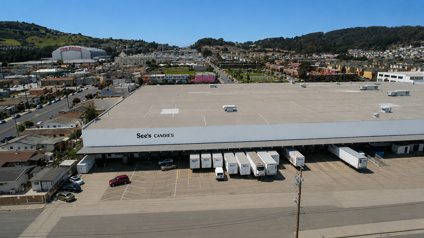A large commercial building with a sign reading 'See's Candies' on the front, surrounded by a parking lot with several parked vehicles and delivery trucks. In the background, there are residential houses, a hill with trees, and a sports arena with a sign that reads 'Cow Palace' in the distance.