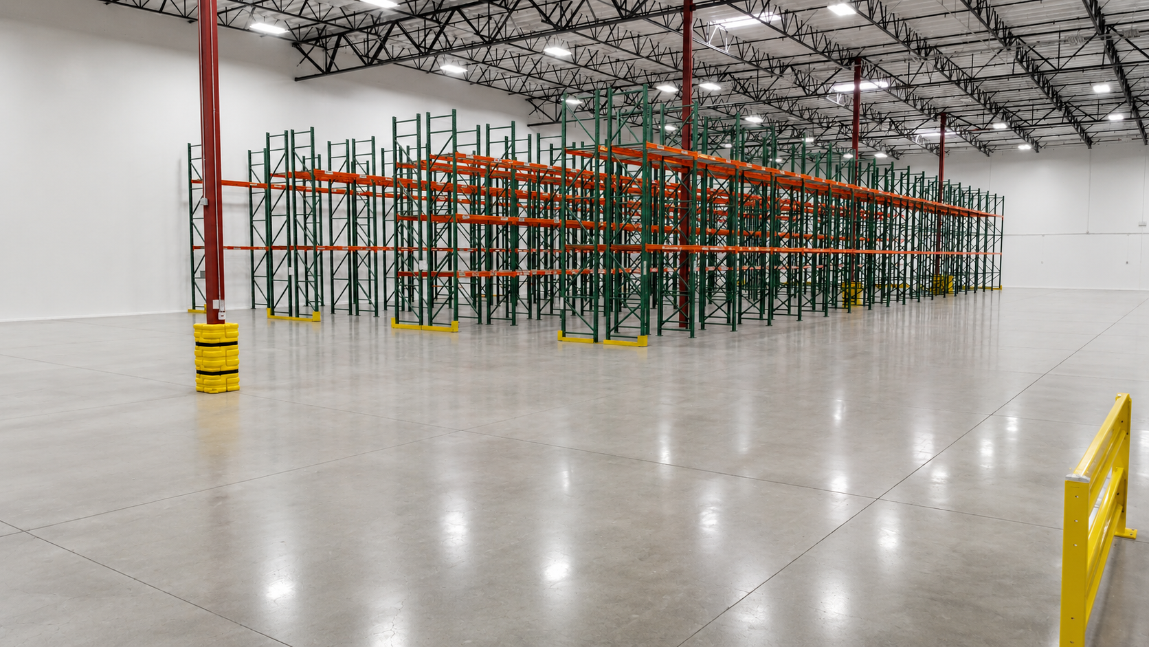 Empty warehouse with green and orange shelving units, yellow safety barriers, and bright lighting.