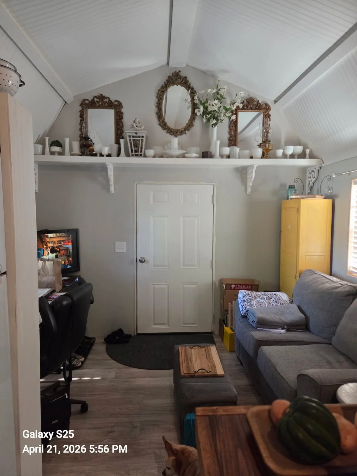 Living room with a gray couch, small ottoman, yellow cabinet, TV in the corner, and decorative shelf with mirrors and vases on the high wall. A door in the center, a window on the right, and a small table with a pumpkin in the foreground.