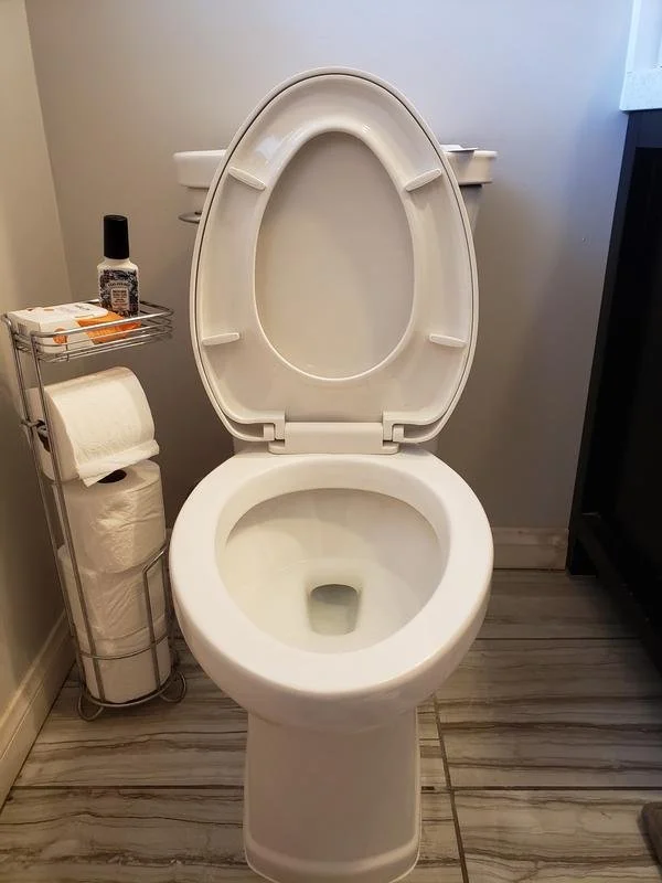 A white toilet with the lid open in a bathroom, next to a small metal shelf holding a bottle of nail polish and a roll of toilet paper on a stand.